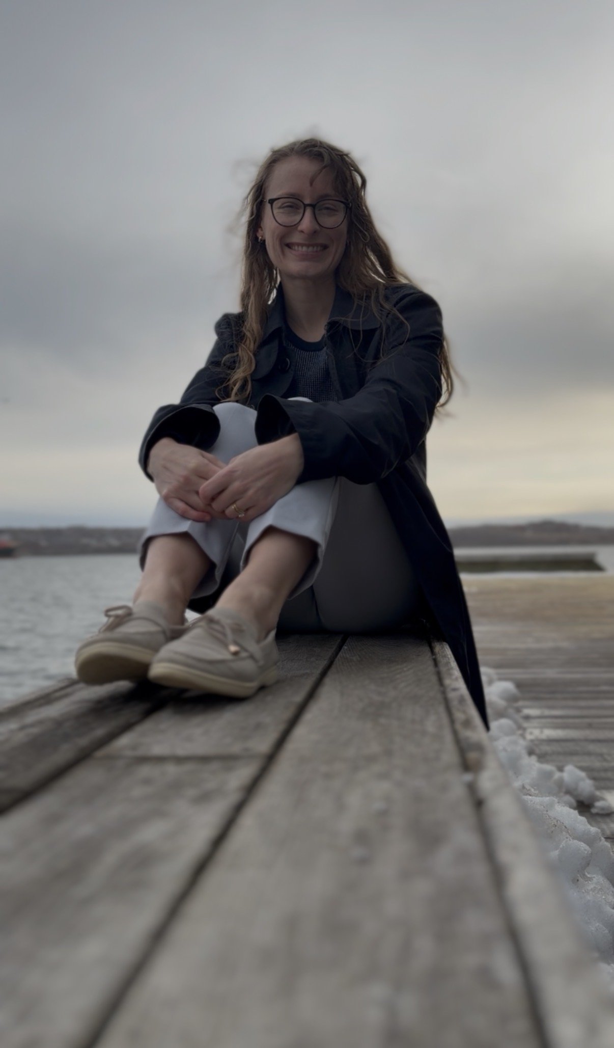 A woman sitting on a wooden dock outdoors near water, smiling at the camera, wearing glasses, a dark jacket, gray pants, and beige shoes.