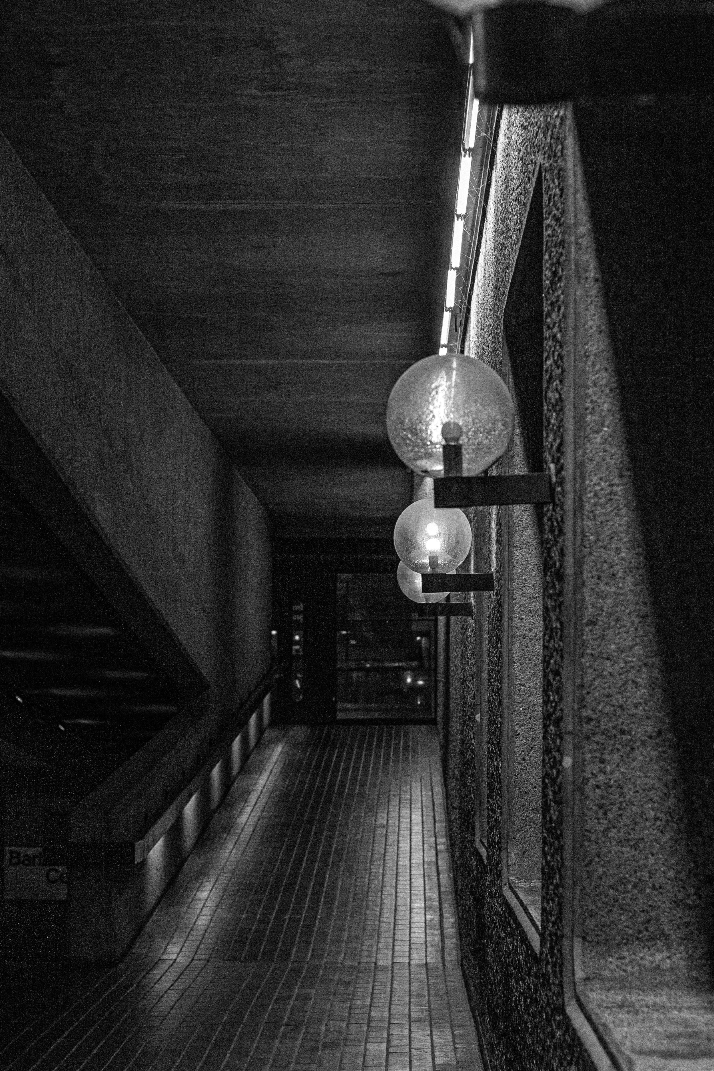 Black and white photograph of an indoor corridor with hanging globe lights on the right wall, and a tiled floor leading to a glass door at the end.