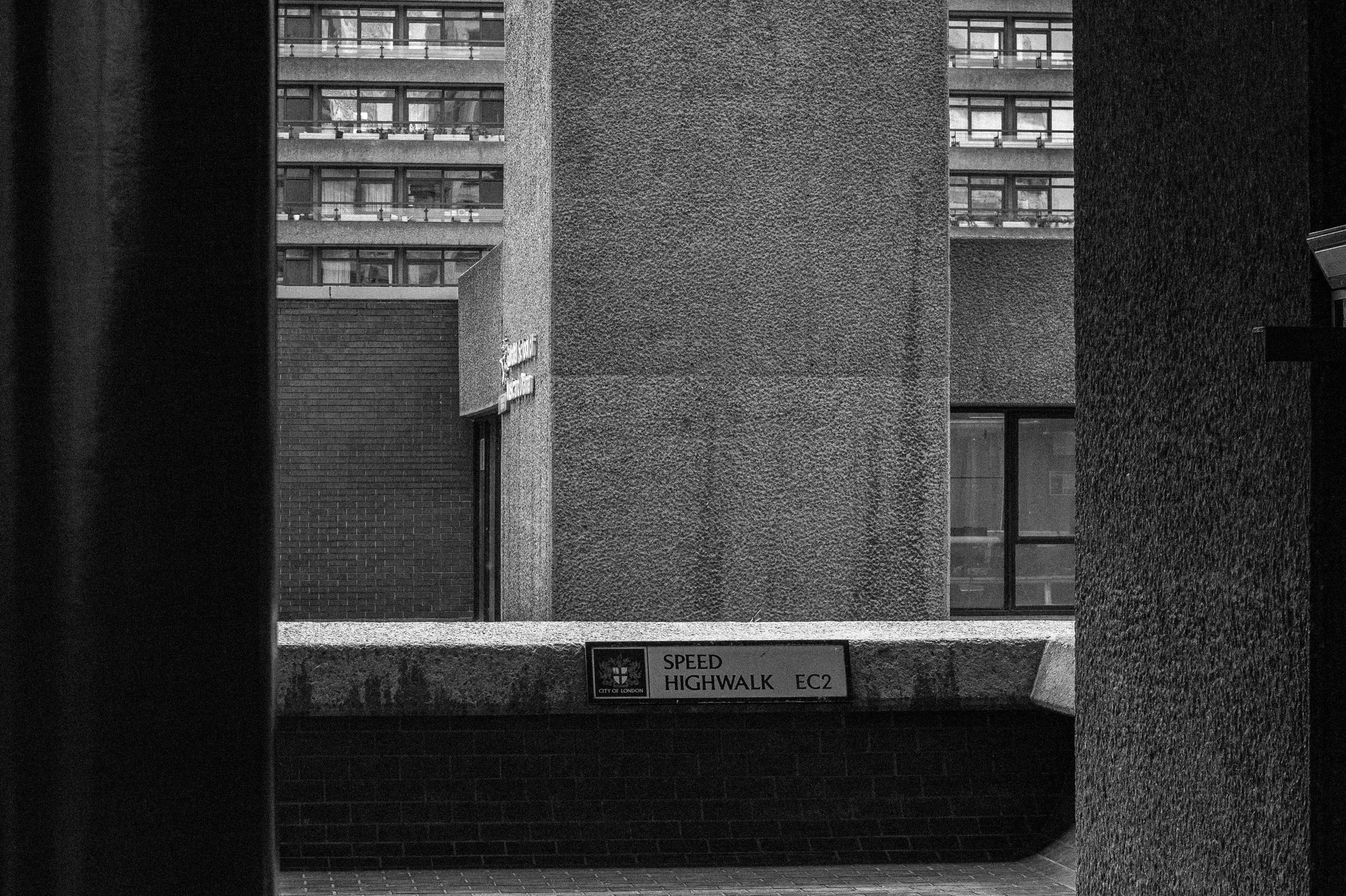 Black and white photo of an urban scene with industrial-style concrete structures and a sign reading 'SPEED HIGHWALK EC2' on a ledge. In the background, there are multi-story buildings with rows of windows and balconies.
