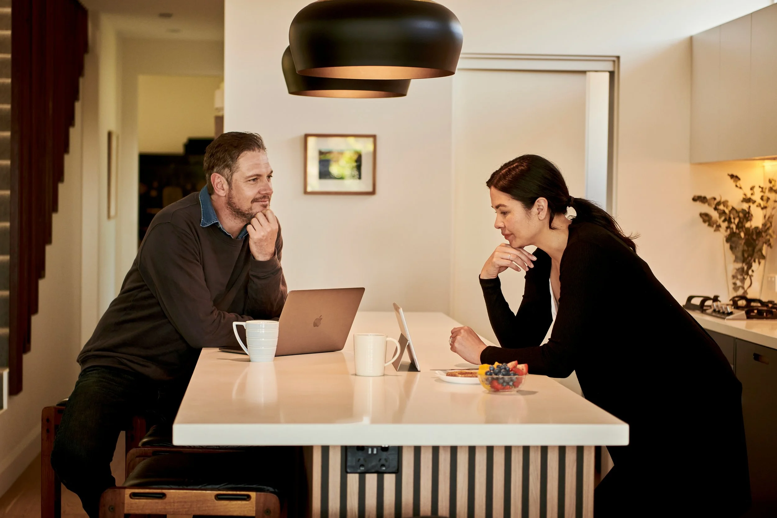 Man and woman having a serious conversation at a kitchen table with laptops and cups.