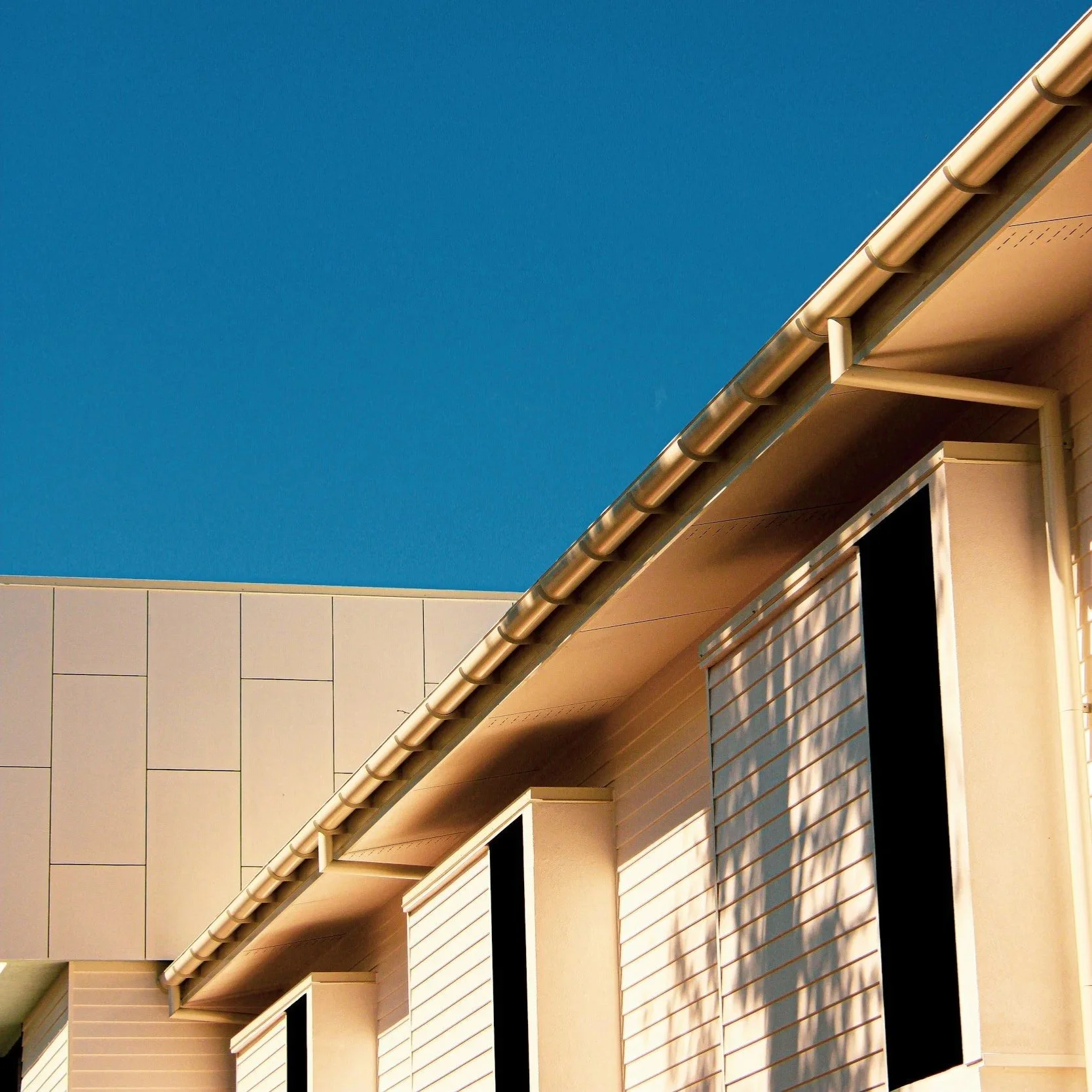 Close-up of a modern building's beige exterior with white shutters on windows, brown roof golden gutters, and blue sky in the background.