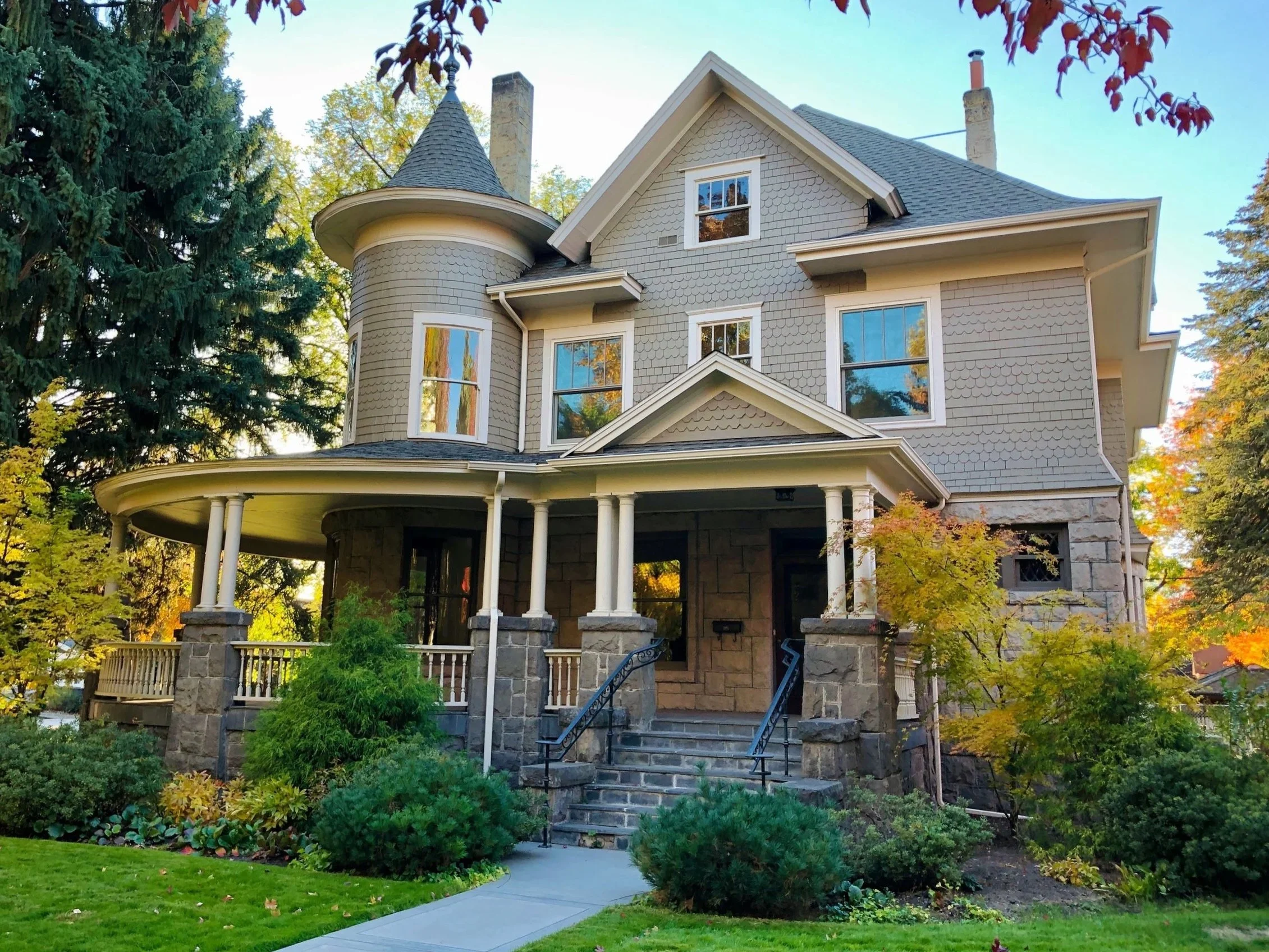 A large Victorian-style house with a turret, stone foundation, and a front porch with columns, surrounded by landscaped garden and trees.