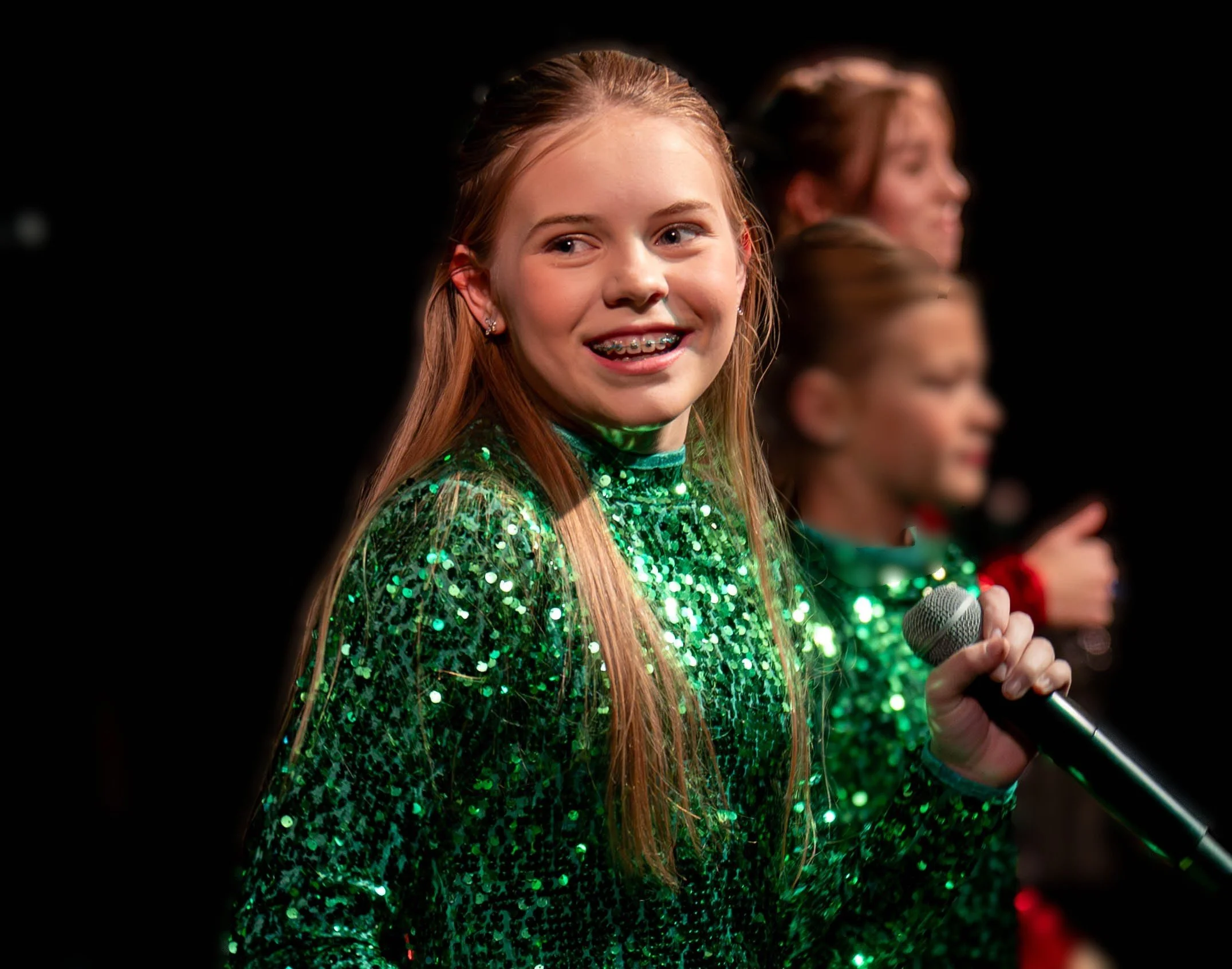 Teen girl in a green dress with a microphone smiling in the direction of the camera.