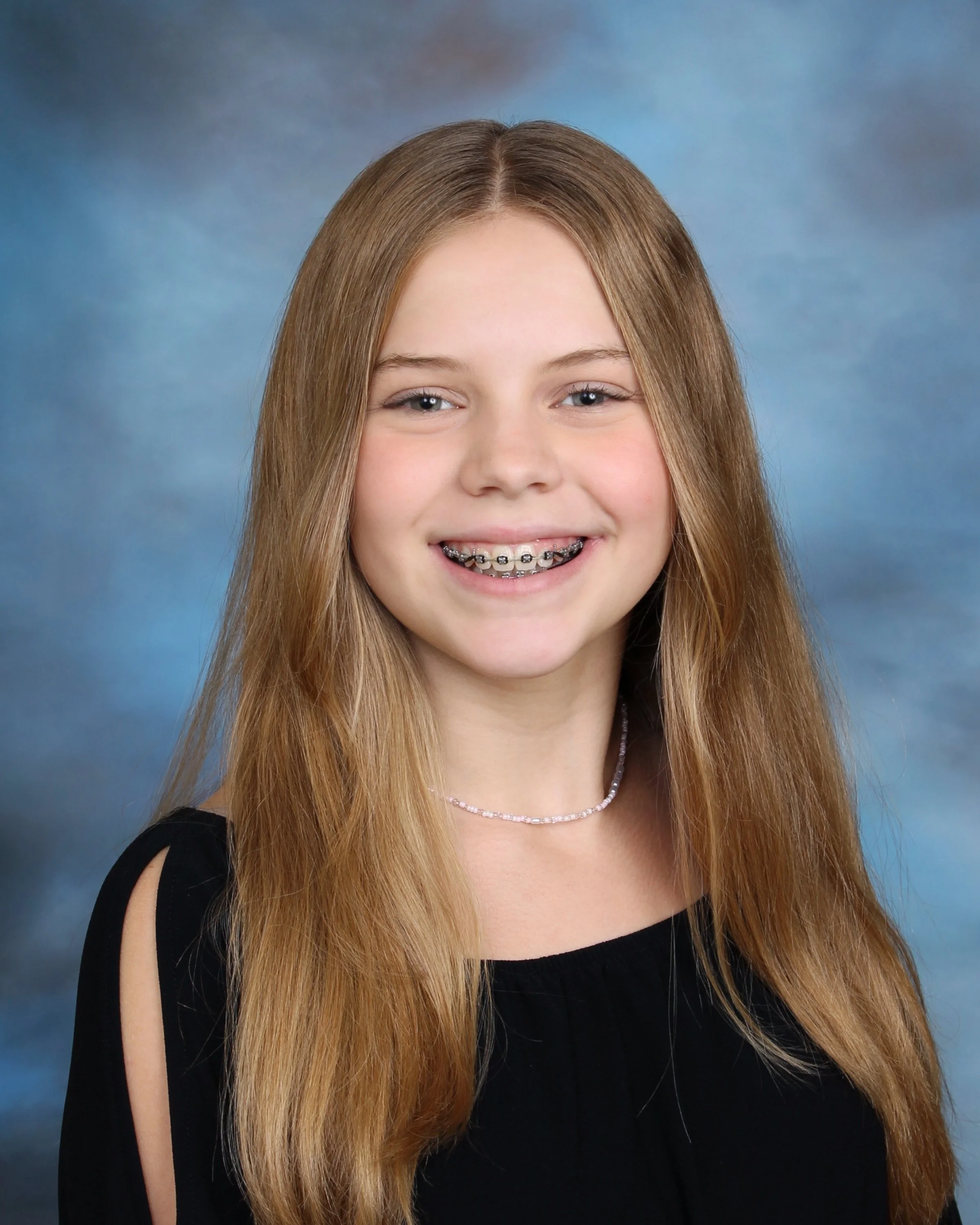 A young girl with long red hair wearing braces and a black top, smiling against a blue and gray background.