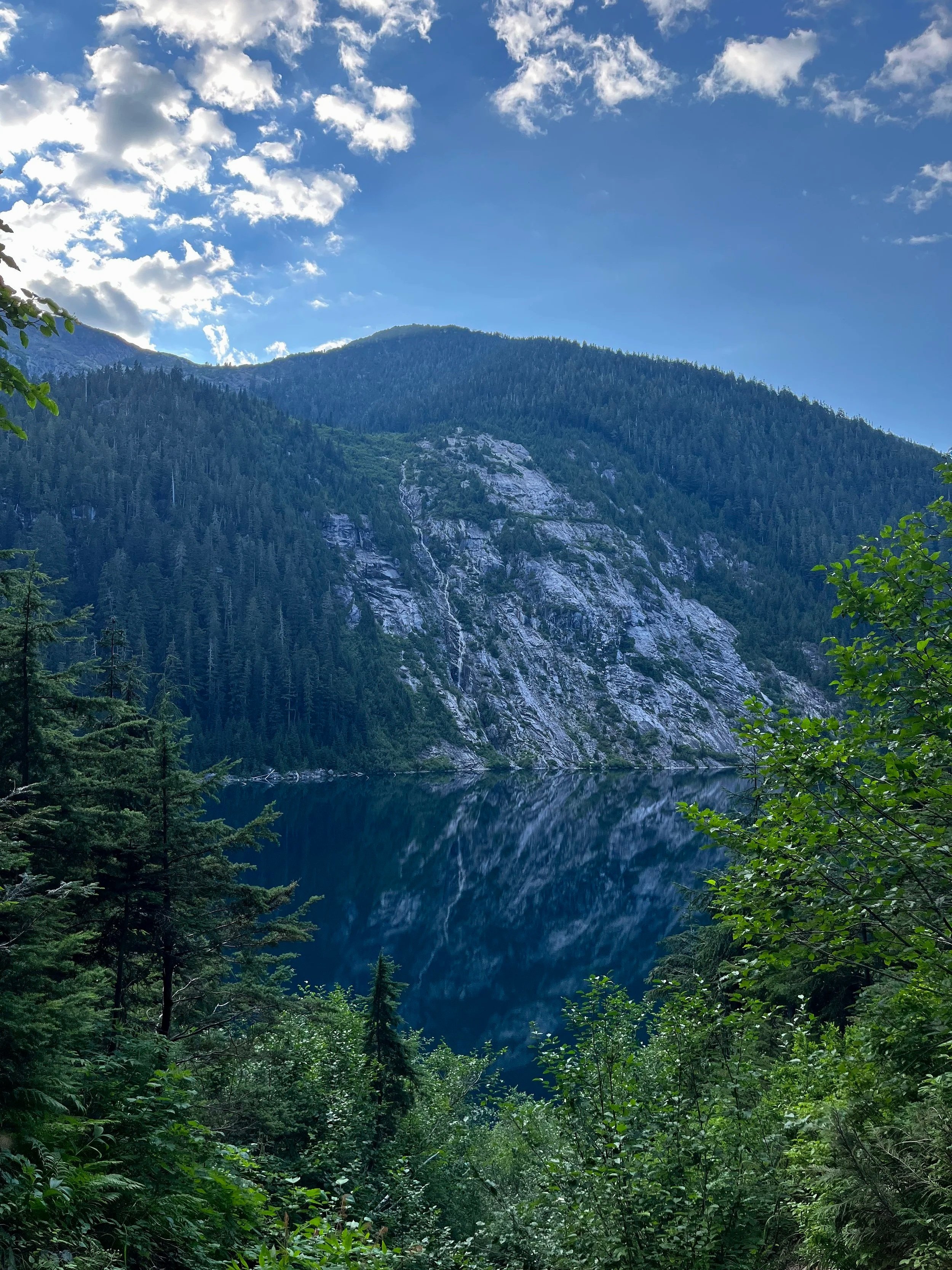 A forested mountain landscape with a lake in the foreground, trees surrounding the water, and a partly cloudy sky above.