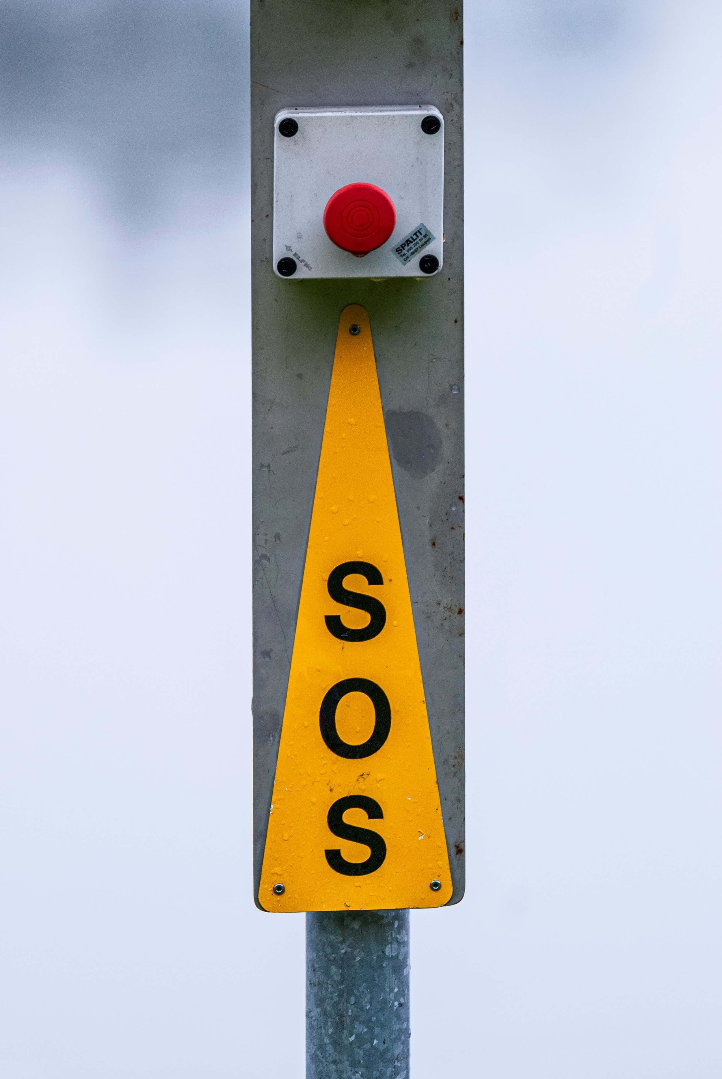 Close-up of a bus SOS emergency button with a red cover, mounted on a metallic pole with a yellow triangle warning sign with the words "SOS" in black letters.