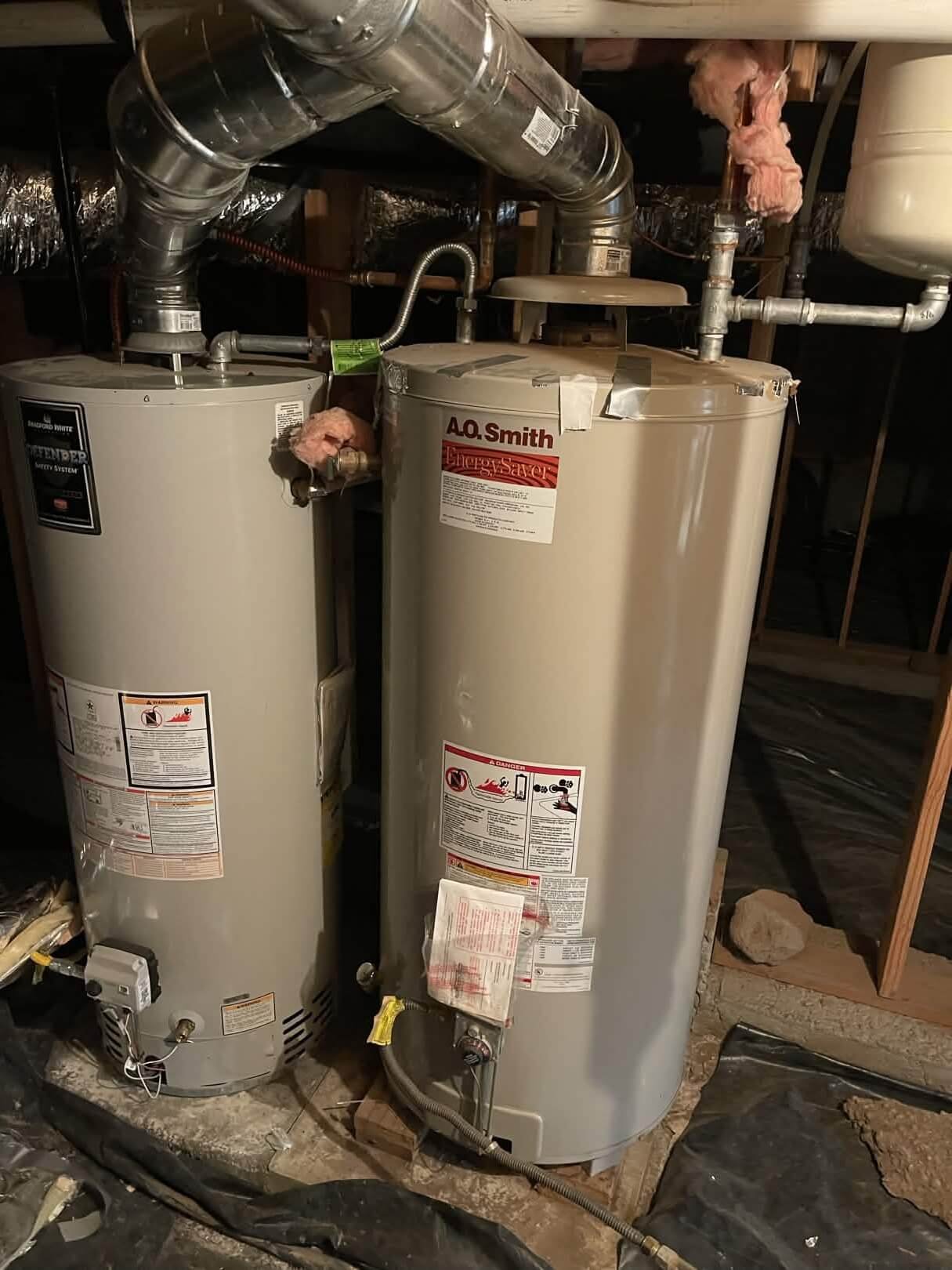 Two traditional cylindrical gas water heaters, a Bradford White and an A.O. Smith Energy Saver, installed side-by-side in a basement with galvanized venting pipes.