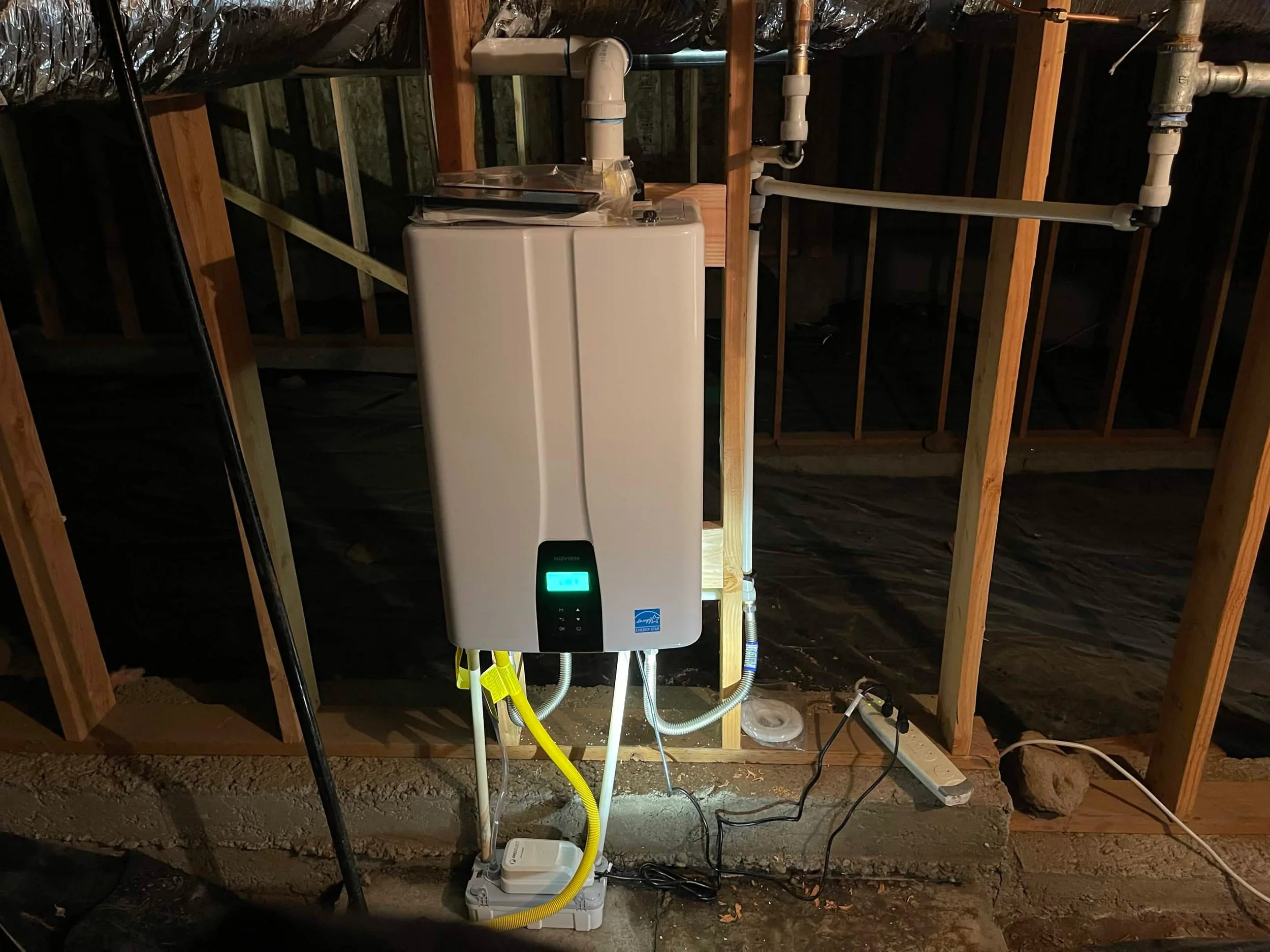 A modern white Navien tankless water heater mounted on wooden wall studs in a crawl space with exposed insulation and a yellow gas line.