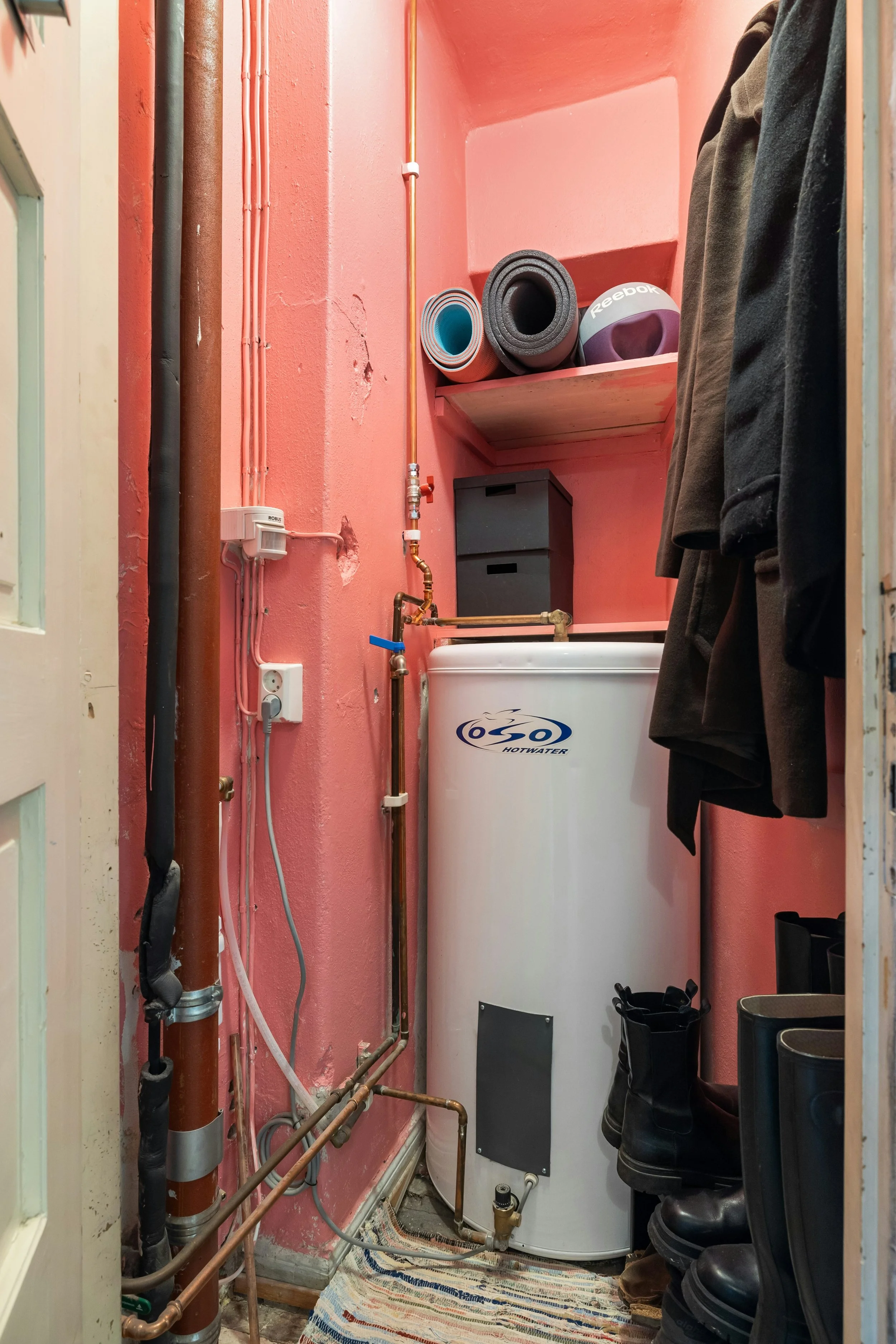 A small utility closet containing a white hot water heater, plumbing pipes, and various clothing items including jackets and boots.