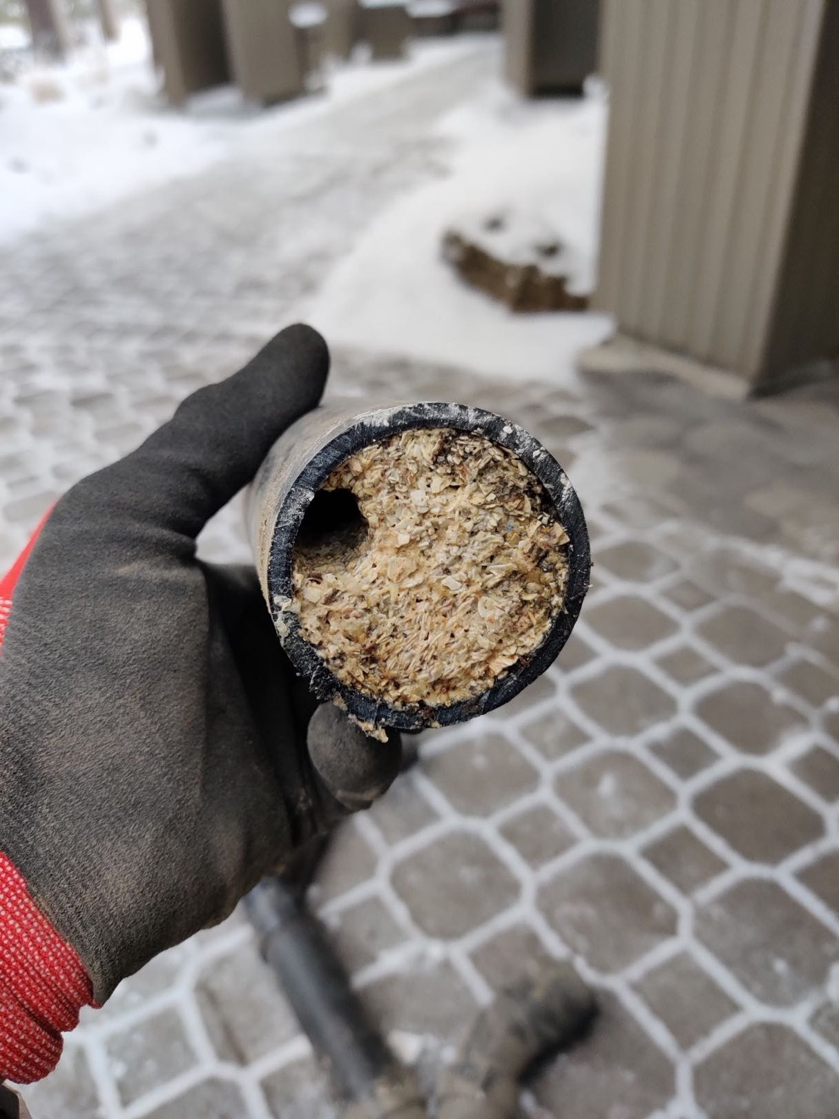 A gloved hand holding up a cross-section of a black pipe that is severely clogged with a thick, fibrous, beige substance. A small opening is visible on the left side of the pipe interior, revealing the extent of the blockage.
