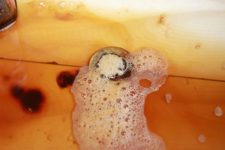 A close-up shot of a white sink filled with murky, amber-colored standing water and soapy bubbles. The rusty-looking liquid is backed up around the metal drain, with dark brown stains visible underwater.