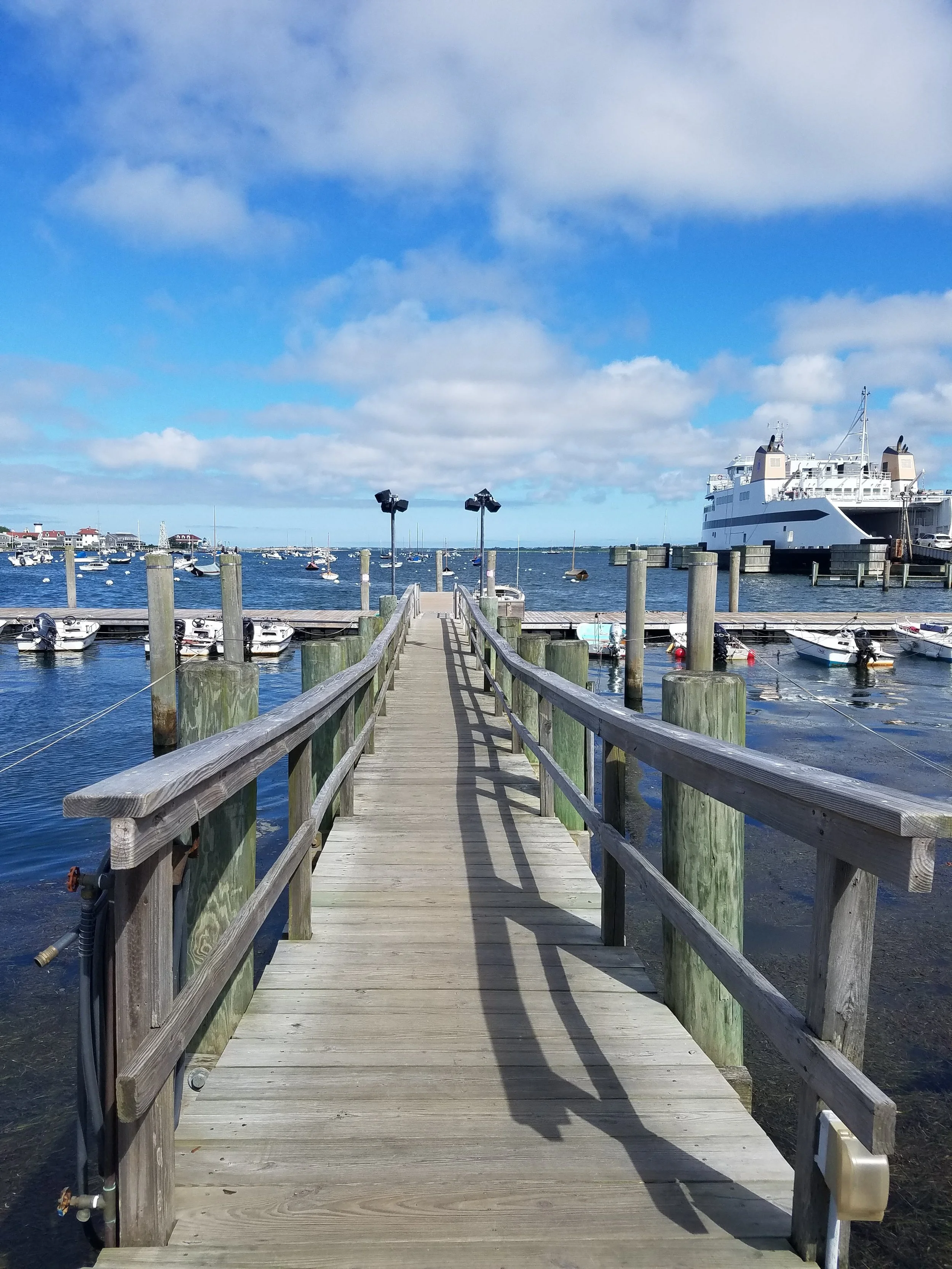Photo of a weathered wooden ramp out over the ocean shore to a dock with a few small boats. A large ferry is in the background and a blue sky with puffy clouds.