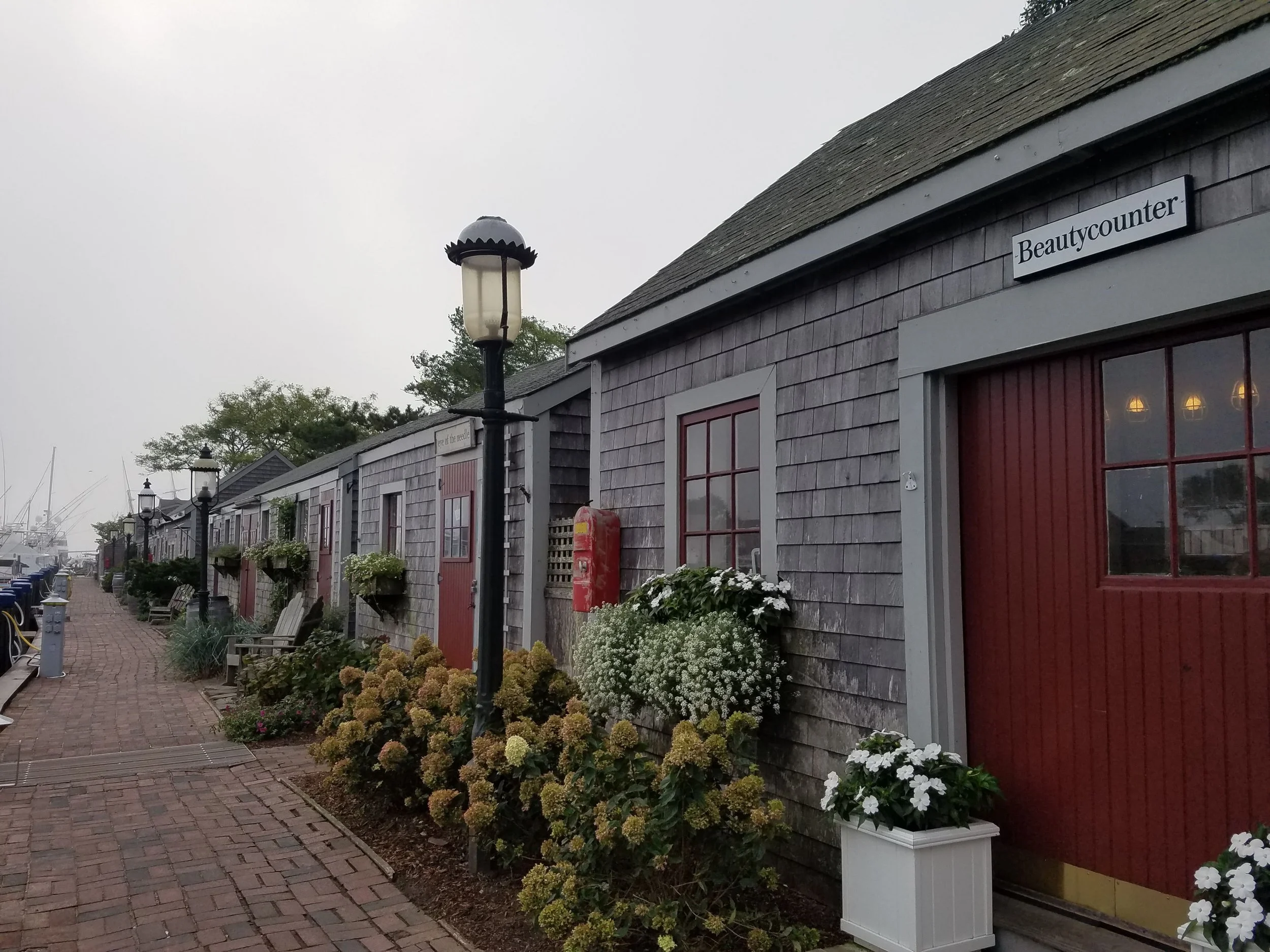 A long series of identical, small cottages with weathered shingles and red doors and red window trim. There are old fashioned street lamps along the way. The cottages are along a brick walkway facing docked boats.