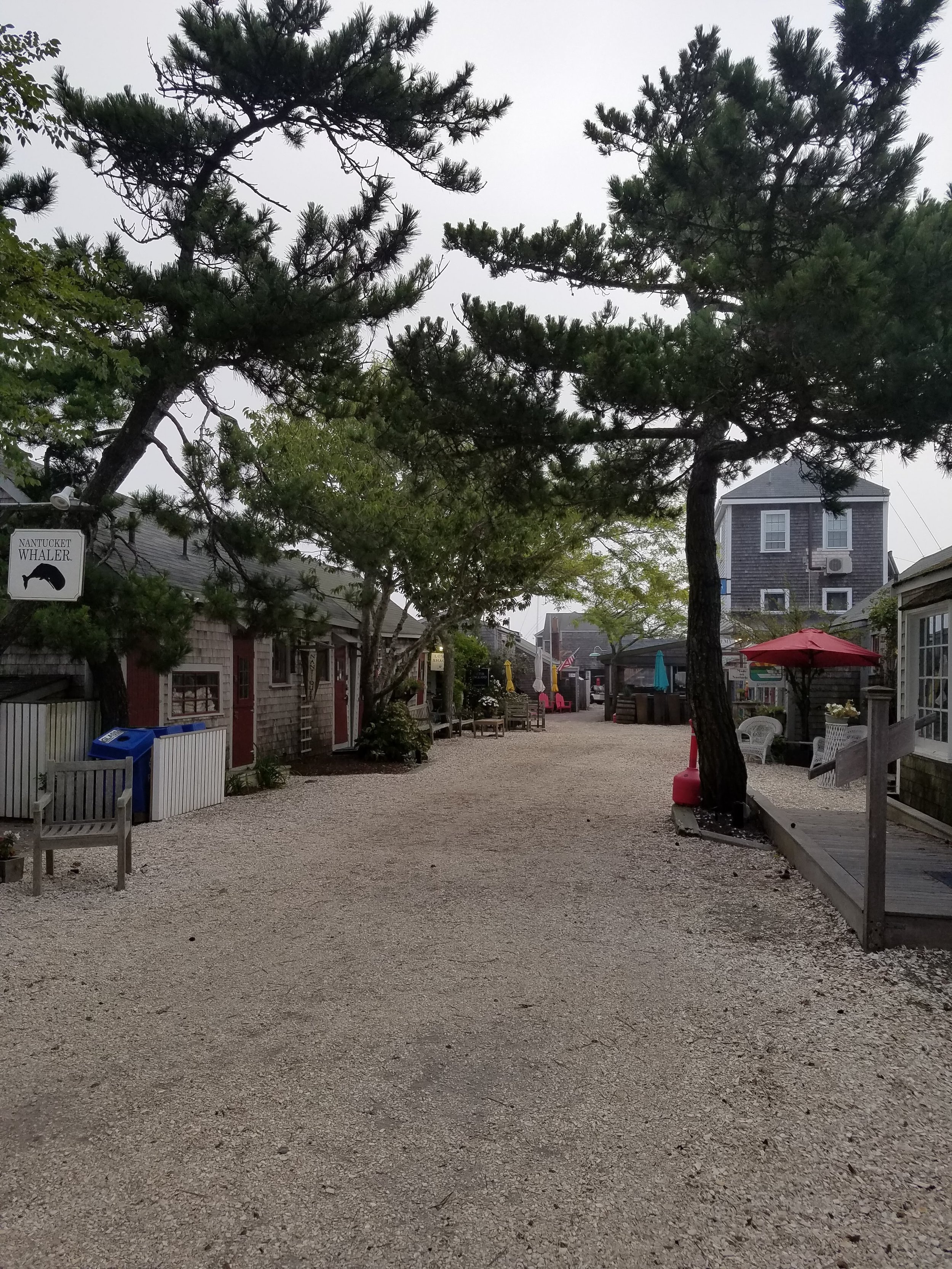 Photo of a pebble and sand road on Nantucket with simple low buildings on the left and a larger old house in the background, all with weathered shingles. There are several trees with plush green branches and a red umbrella over a sitting area.