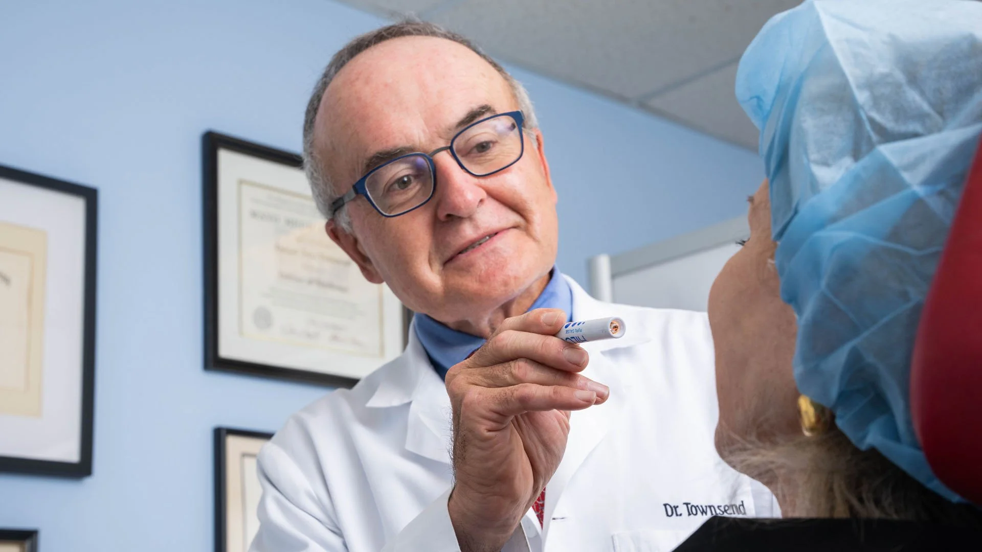 Close up photo of Dr. Daniel Townsend as he uses a small flashlight to examine a patient. He is wearing glasses and has a blue shirt under his white lab coat. There are framed diplomas on the wall in the background.