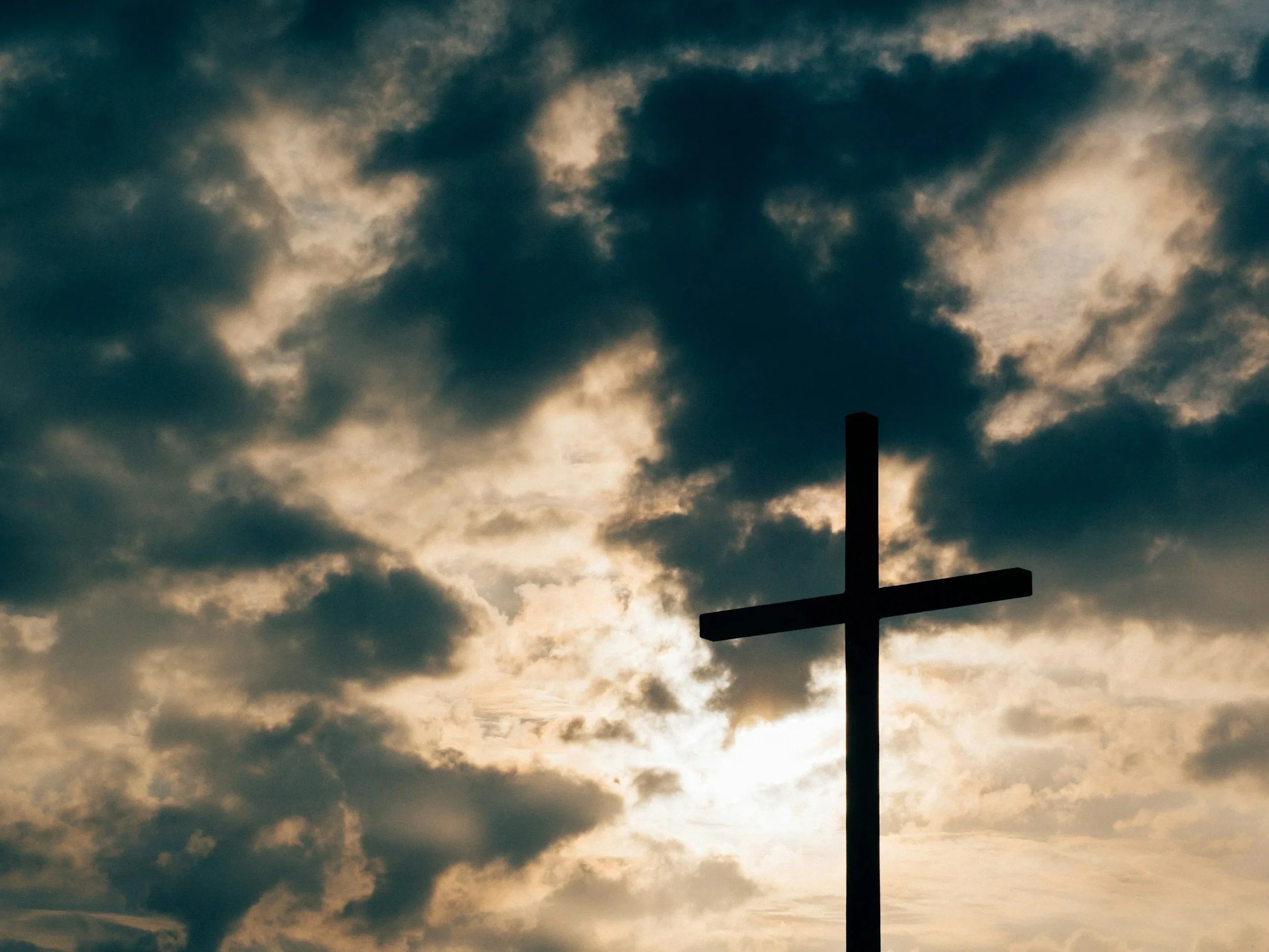 Silhouette of a Christian cross against a cloudy sky with sunlight breaking through.