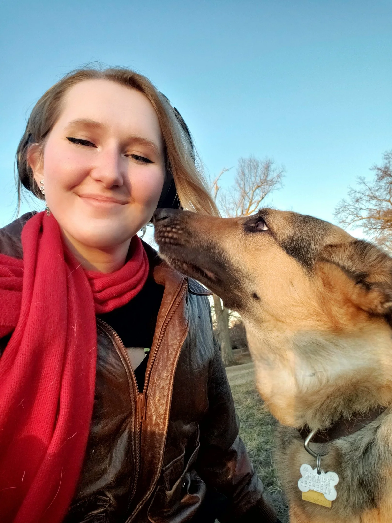 A young woman with blonde hair, wearing a black jacket and red scarf, smiling outdoors with a happy German shepherd looking at her sweetly.