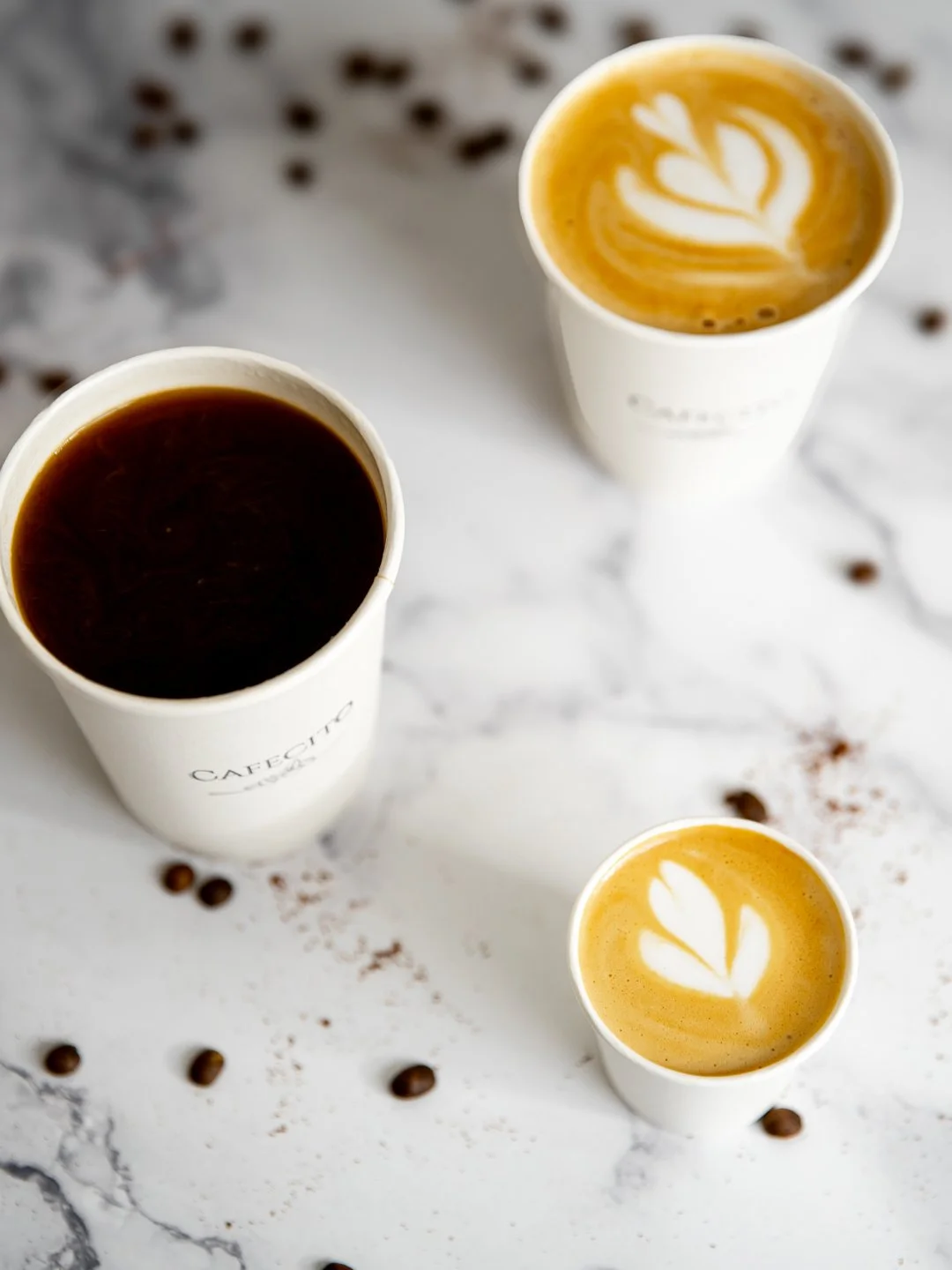 Three cups of coffee on a white marble surface, with scattered coffee beans. Two cups contain latte with leaf-shaped foam art, and one cup contains black coffee.
