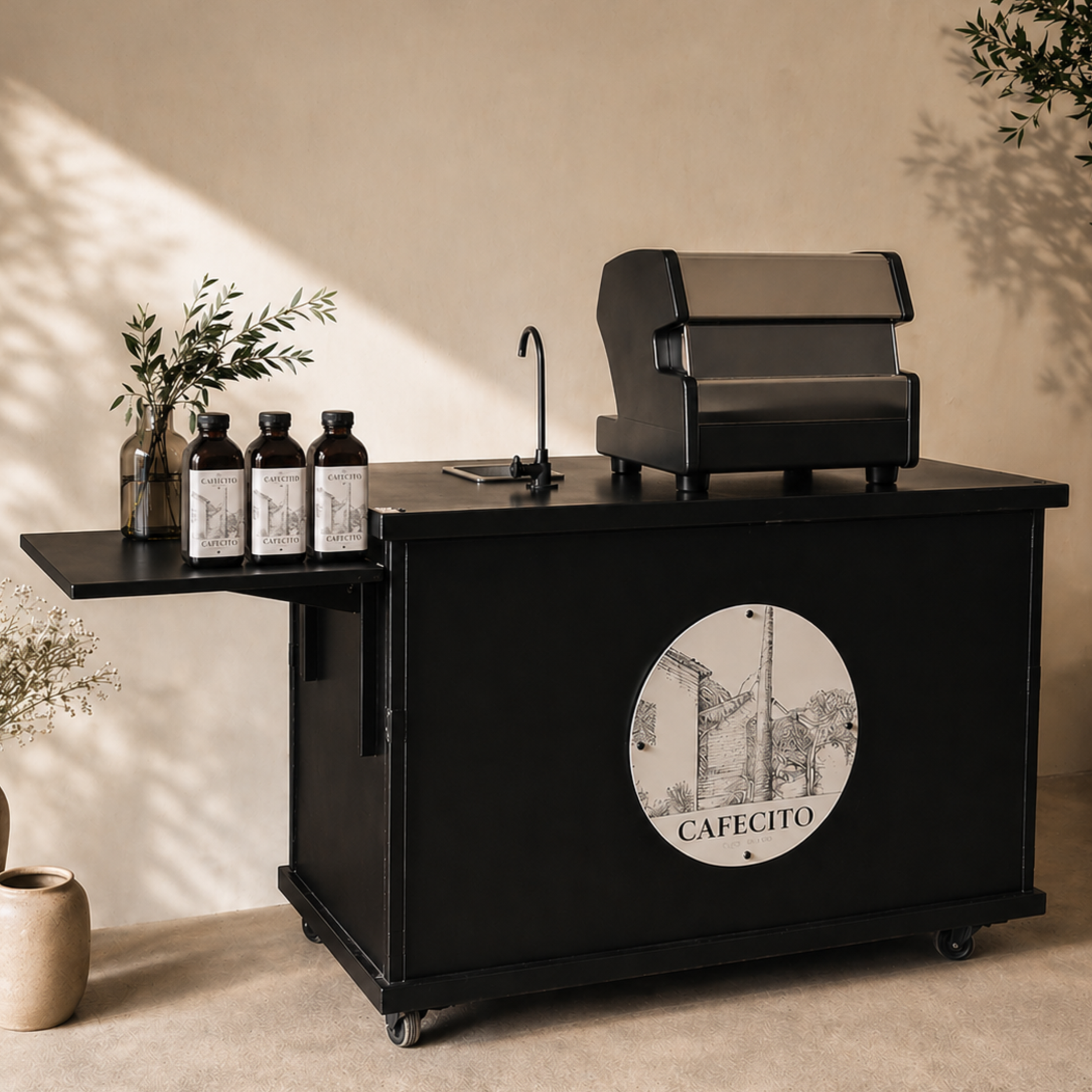 Black mobile cart with a logo reading 'CAFECITO' with an illustration of a windmill; on top, there are three brown bottles with white labels, a glass jar with green leaves, a black faucet, and a black top appliance, with a beige wall and floor in the background.