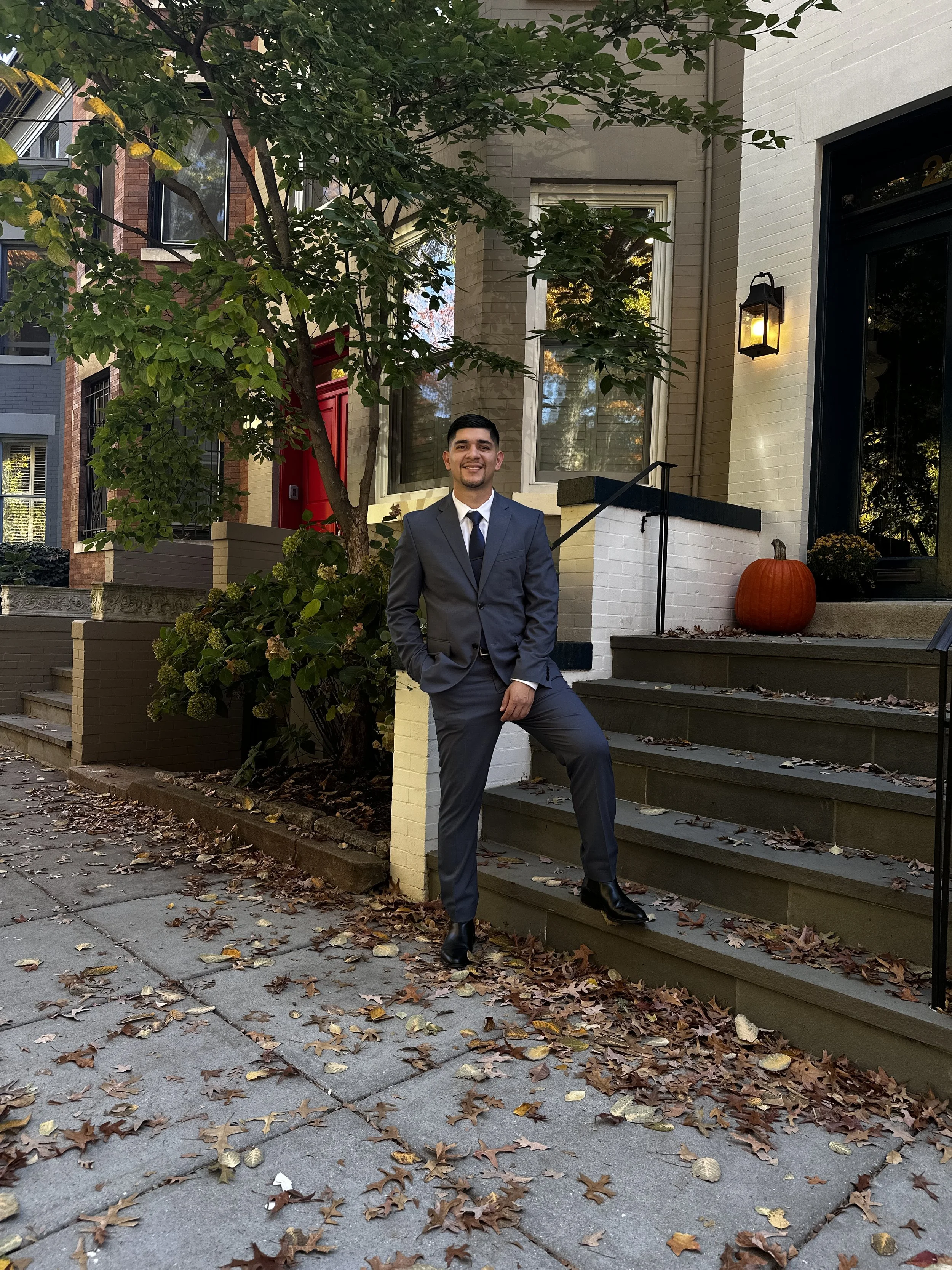 A young man in a suit and tie is standing outdoors next to stairs leading to a house, with a pumpkin on the landing. Autumn leaves are scattered on the sidewalk and steps. The house has a lit outdoor lantern, large windows, and a tree with green leaves partially shading the scene.