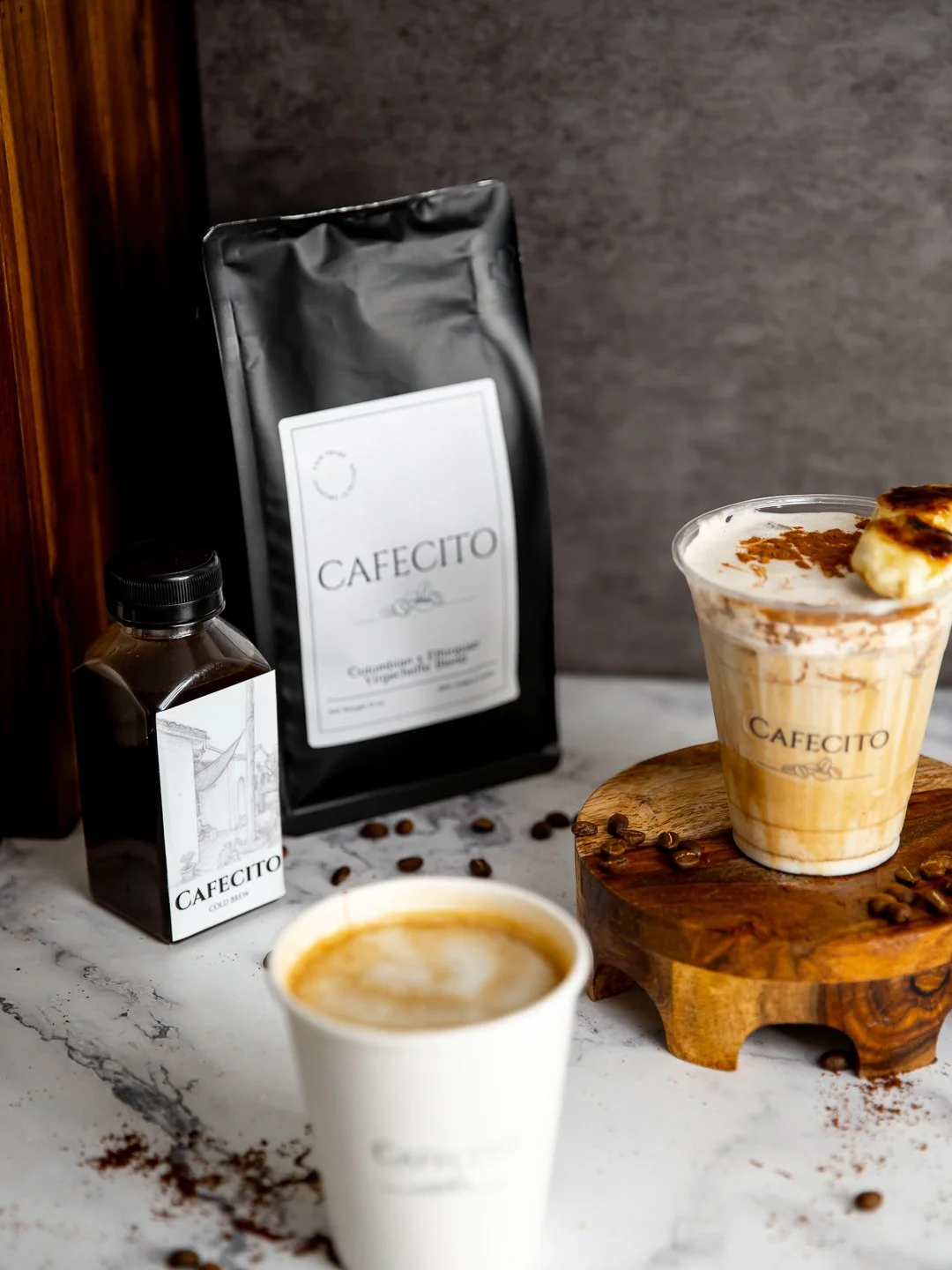 Two coffee drinks, on a wooden stand, surrounded by coffee beans. Behind them are bags of CAFECITO coffee and a bottled coldbrew, all on a marble surface.