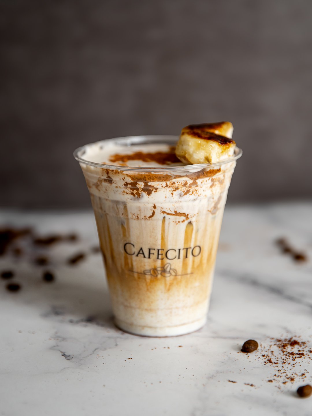 A cup of iced coffee topped with cold foam and cocoa powder, garnished with a caramelized banana, also know as the Banana Bliss Iced Latte.