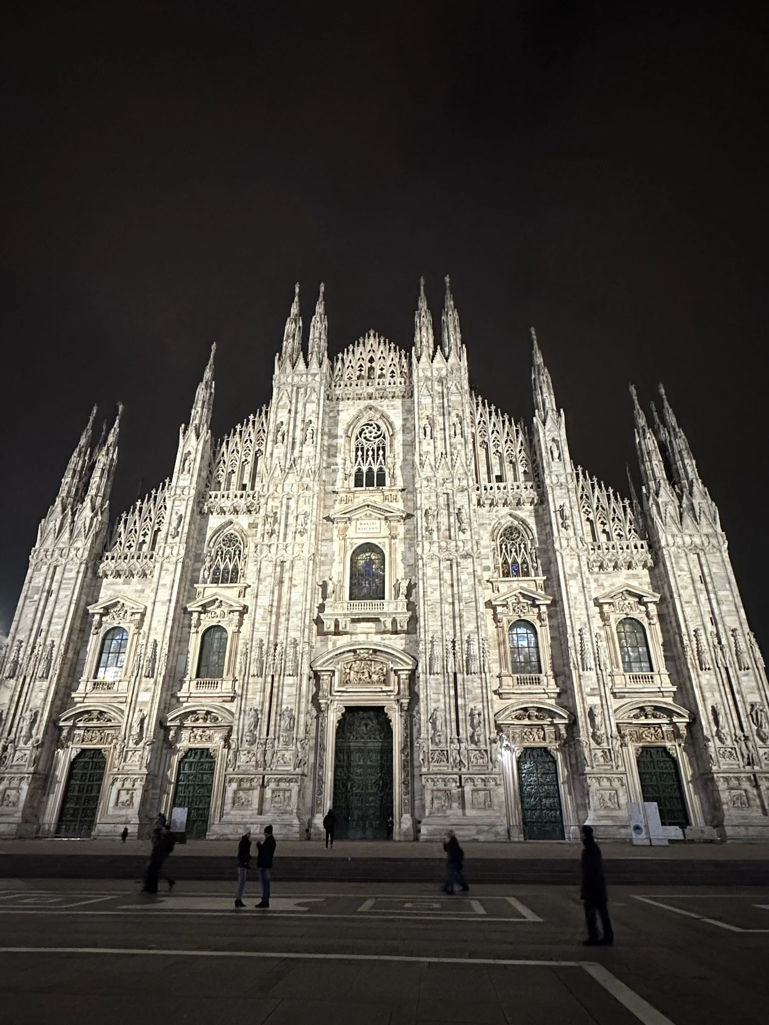 A nighttime view of the illuminated Milan Cathedral (Duomo di Milano) with people standing and walking in front.
