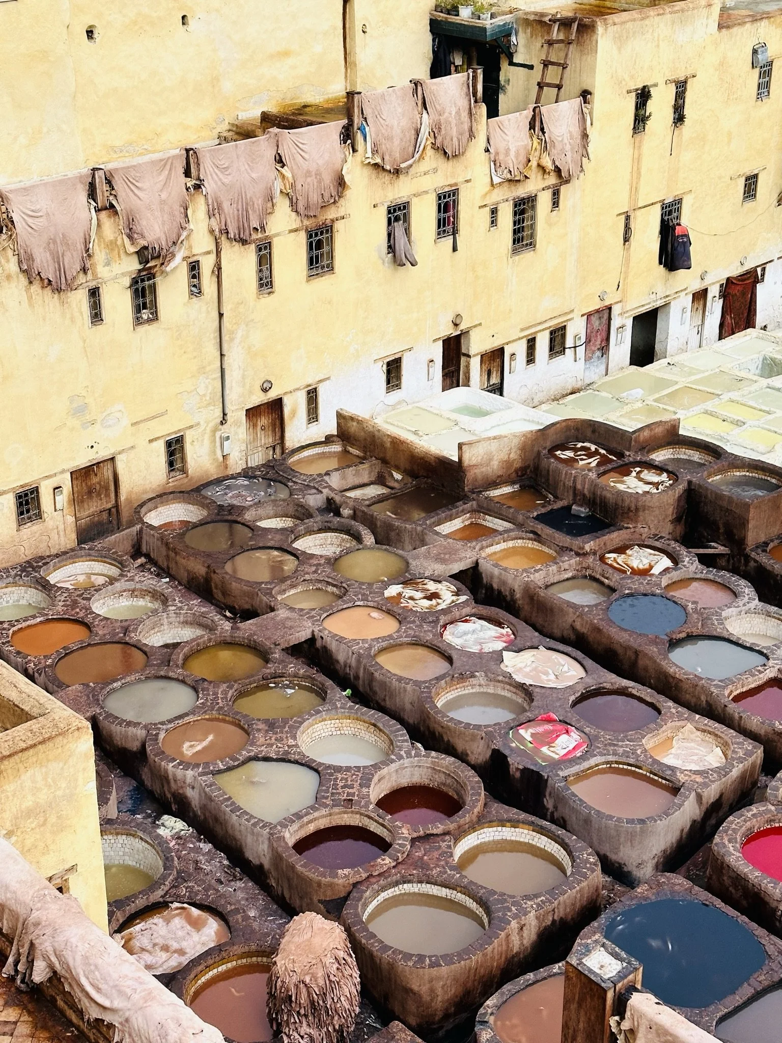 A traditional Moroccan tanner's courtyard with numerous dyeing vats filled with colorful liquids for leather tanning, adjacent to a yellow building with cloth and laundry hanging to dry.