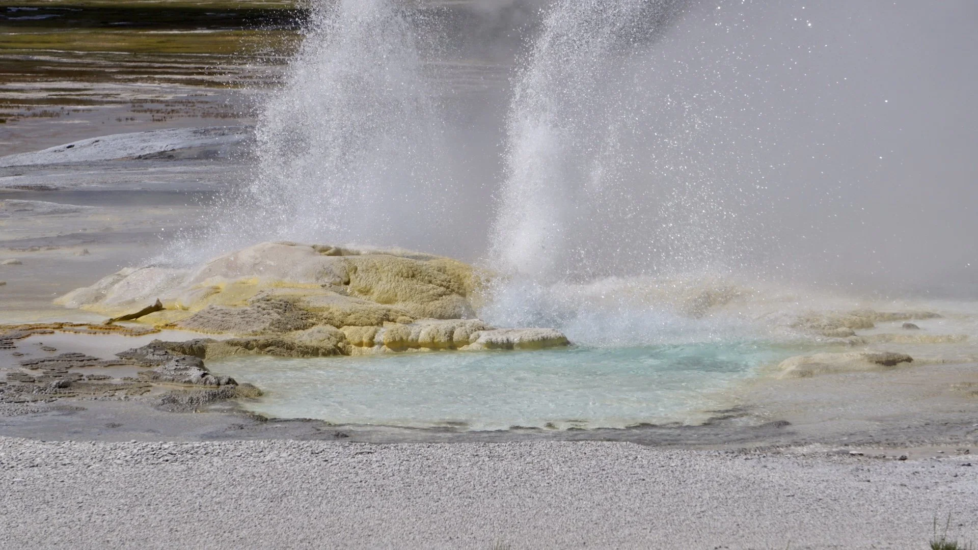 Geyser erupting in hot spring, releasing steam and water into the air, with mineral deposits around the bubbling water in a natural landscape.
