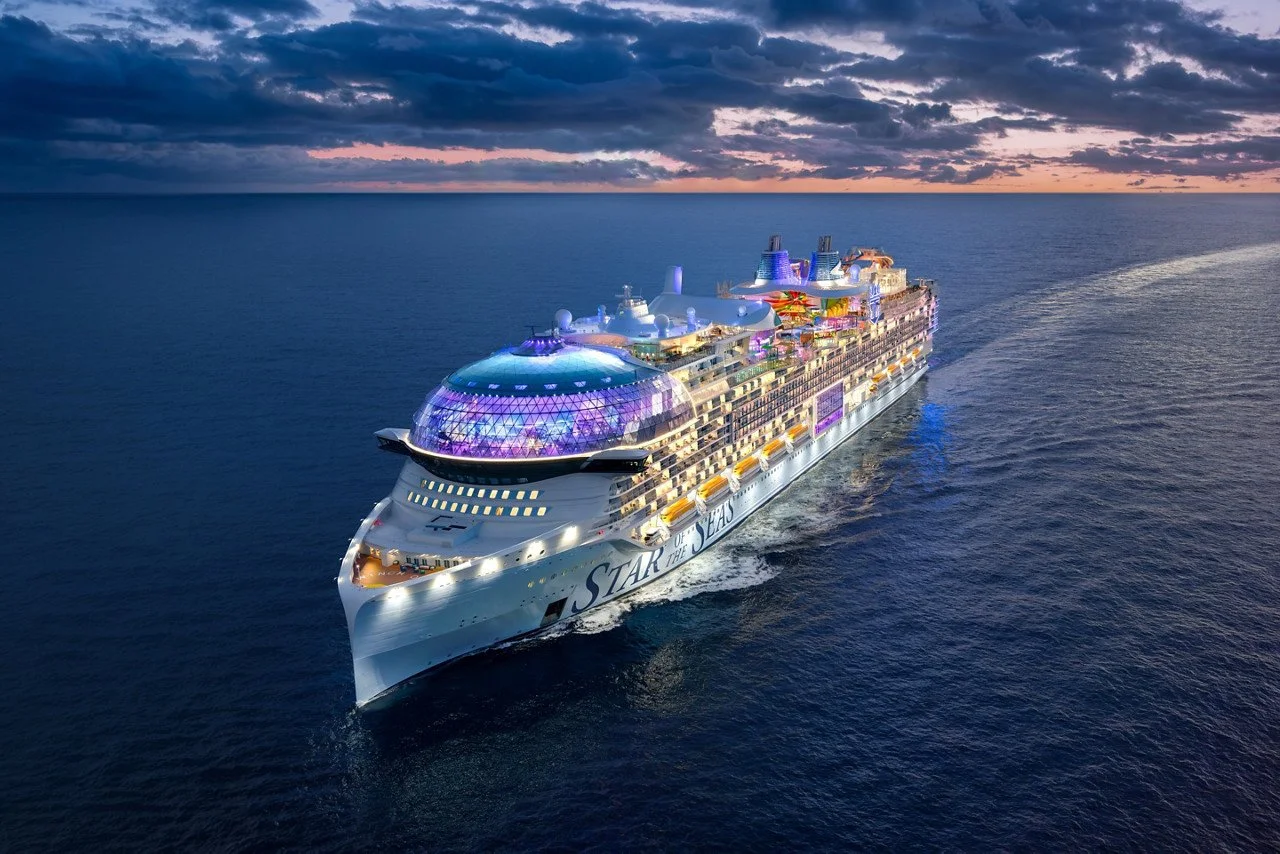 A large, illuminated cruise ship sailing on the ocean at sunset, with a cloudy sky in the background.