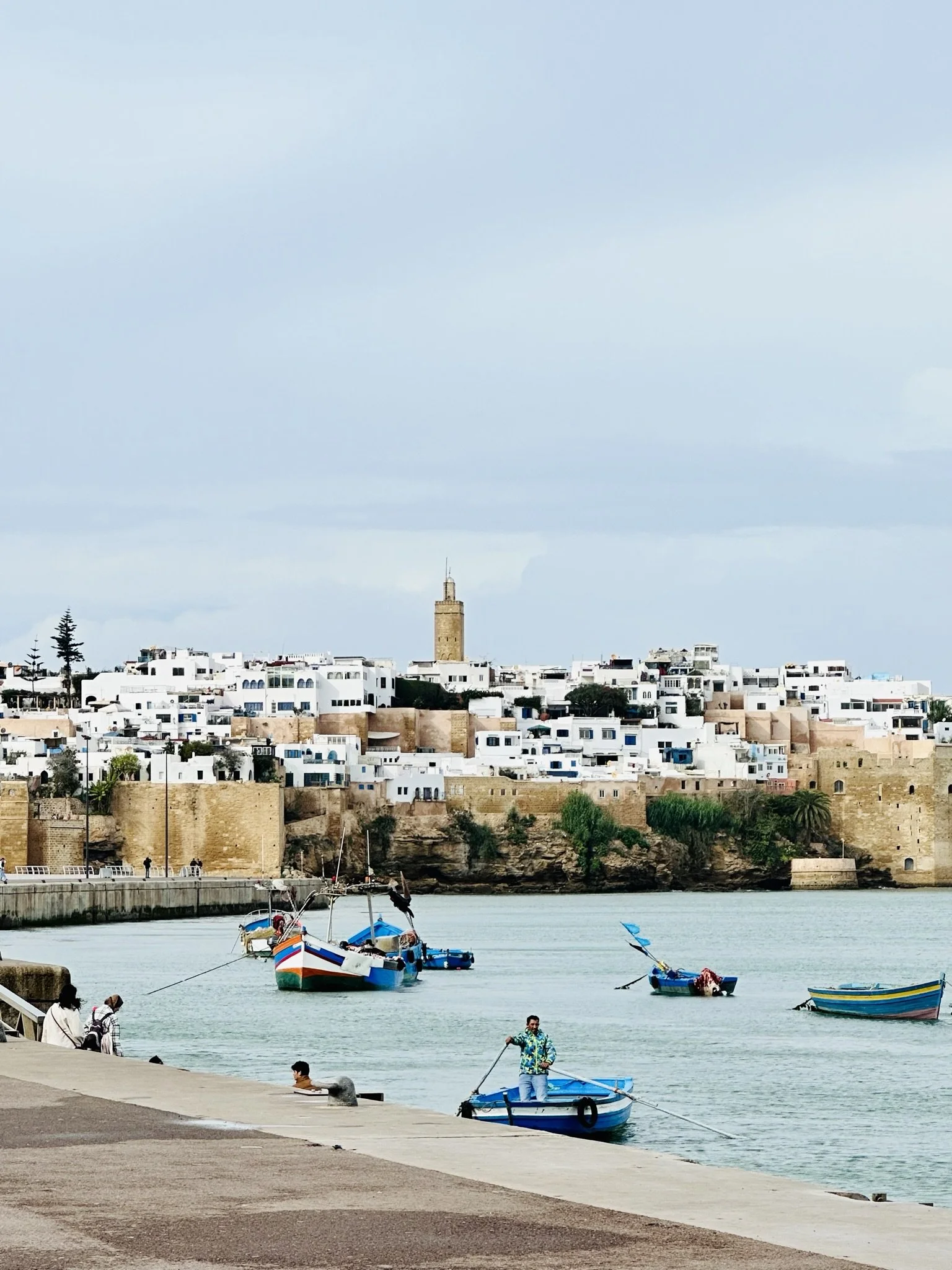 A coastal city with white buildings and a castle-like tower, boats in the water, and people on the dock.