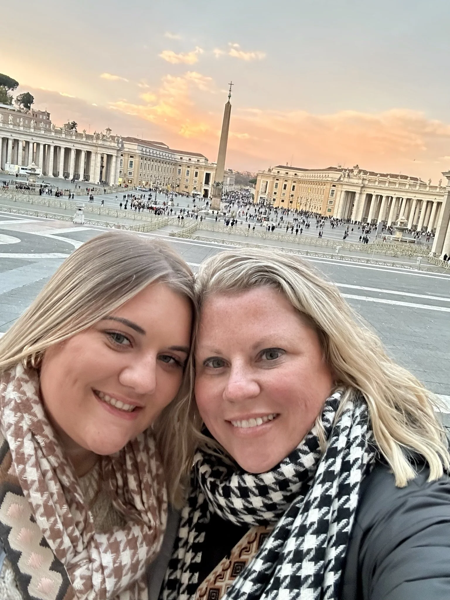 Two women taking a selfie in St. Peter's Square, Vatican City, with the obelisk and classical buildings at sunset in the background.