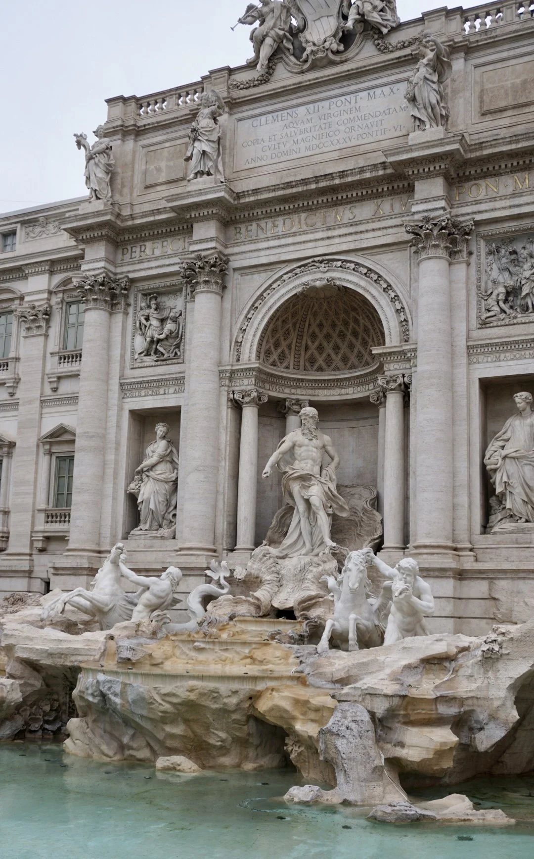 Statues of historical figures and mythological figures atop an ornate building facade, with a prominent fountain with sculptures of mythological creatures in front.