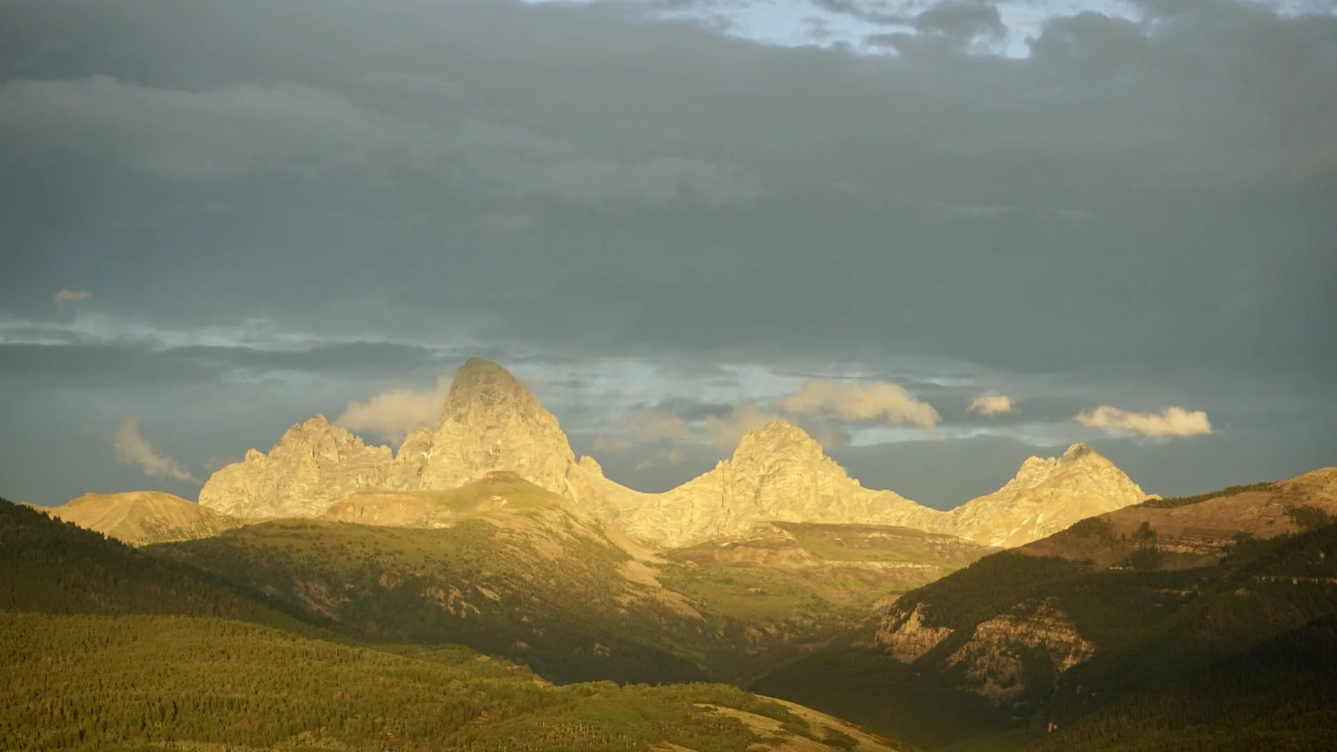 Sunset lighting on rugged mountain peaks with green, forested slopes in the foreground and dark, cloudy sky overhead.