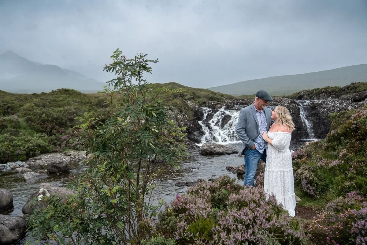 A couple standing close together near a small waterfall in a lush, green, mountainous landscape, with purple flowers and rocks around them, under a cloudy sky.