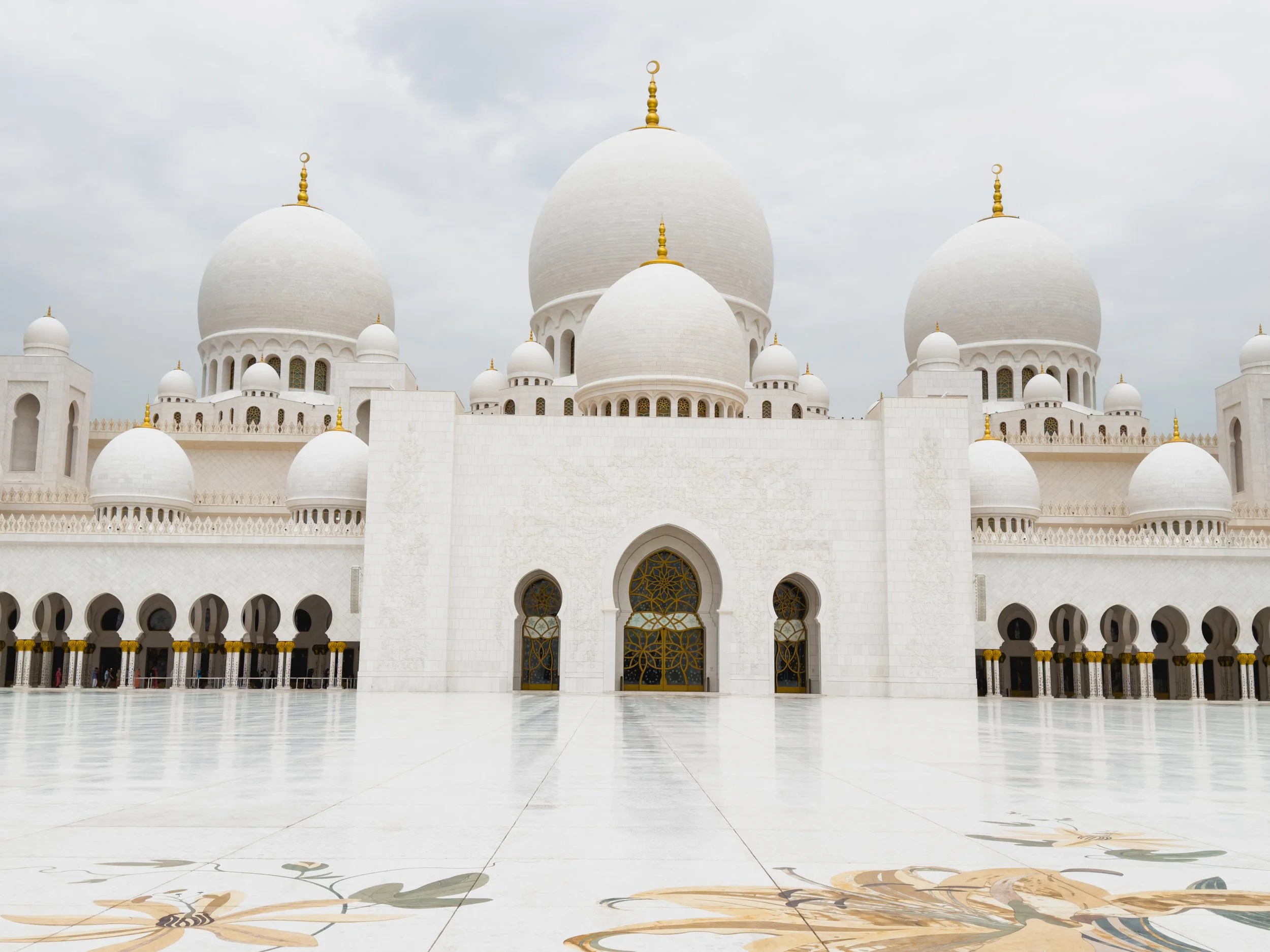 White mosque with multiple domes, arches, and decorative gold details, set against a cloudy sky.