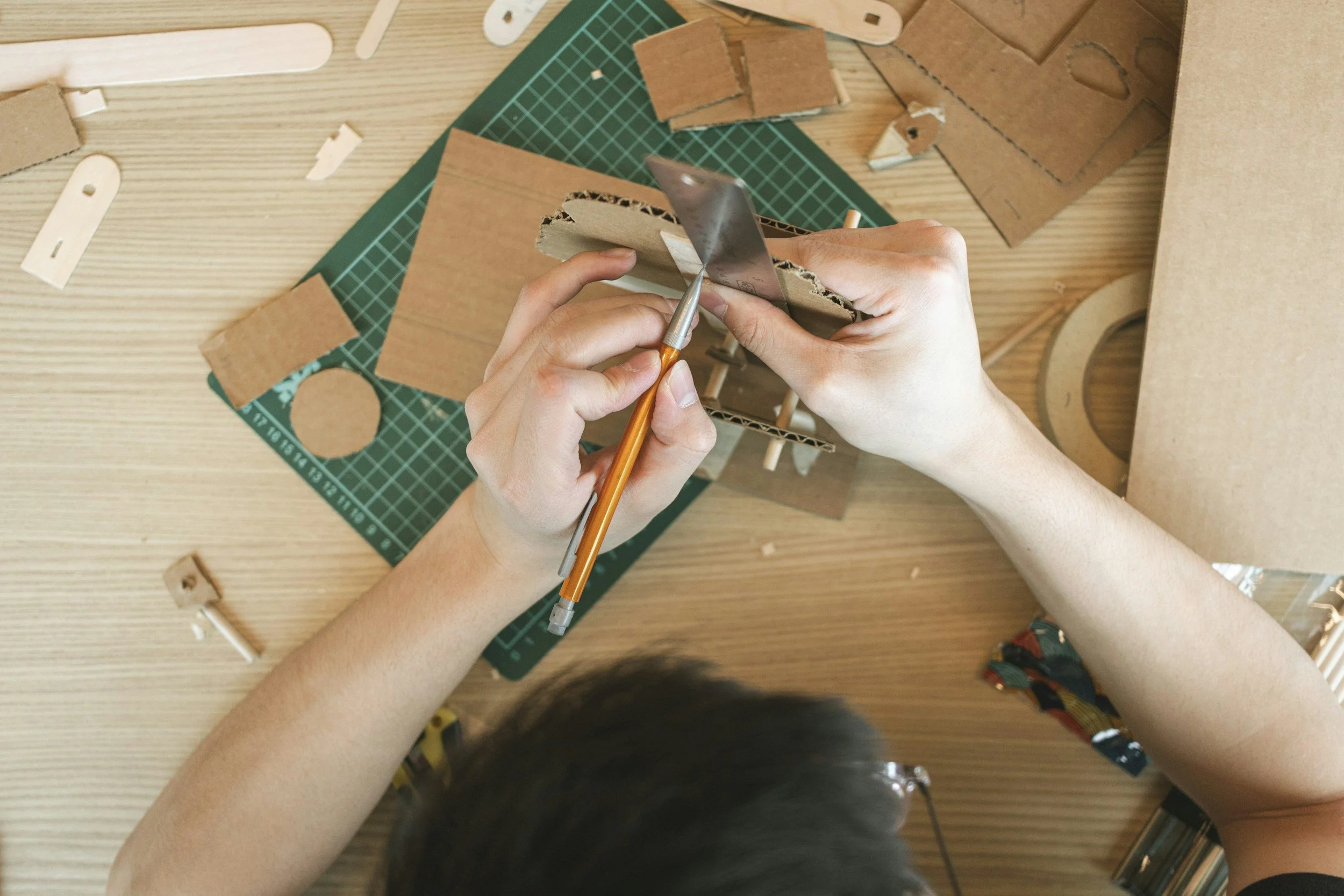 A person assembling a cardboard model with a utility knife, surrounded by various pieces of cardboard on a work surface.
