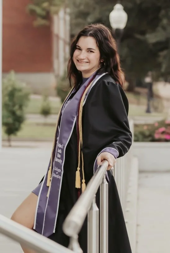 A young woman in graduation gown and honor cords smiling outdoors on campus, leaning on a railing.