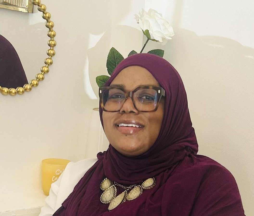 A woman wearing glasses and a maroon hijab, surrounded by decorative items including a gold beaded mirror, a white flower, and a yellow cup in a cozy, well-decorated room.