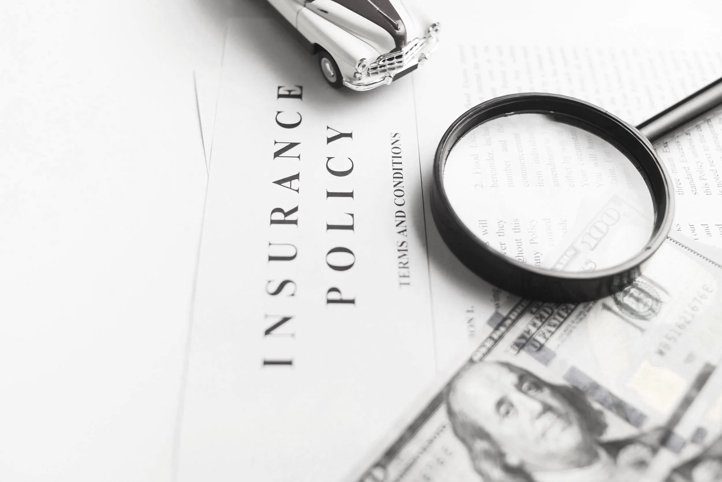A small toy car, a magnifying glass, and a 100-dollar bill placed on a document titled "Insurance Policies" in black and white.