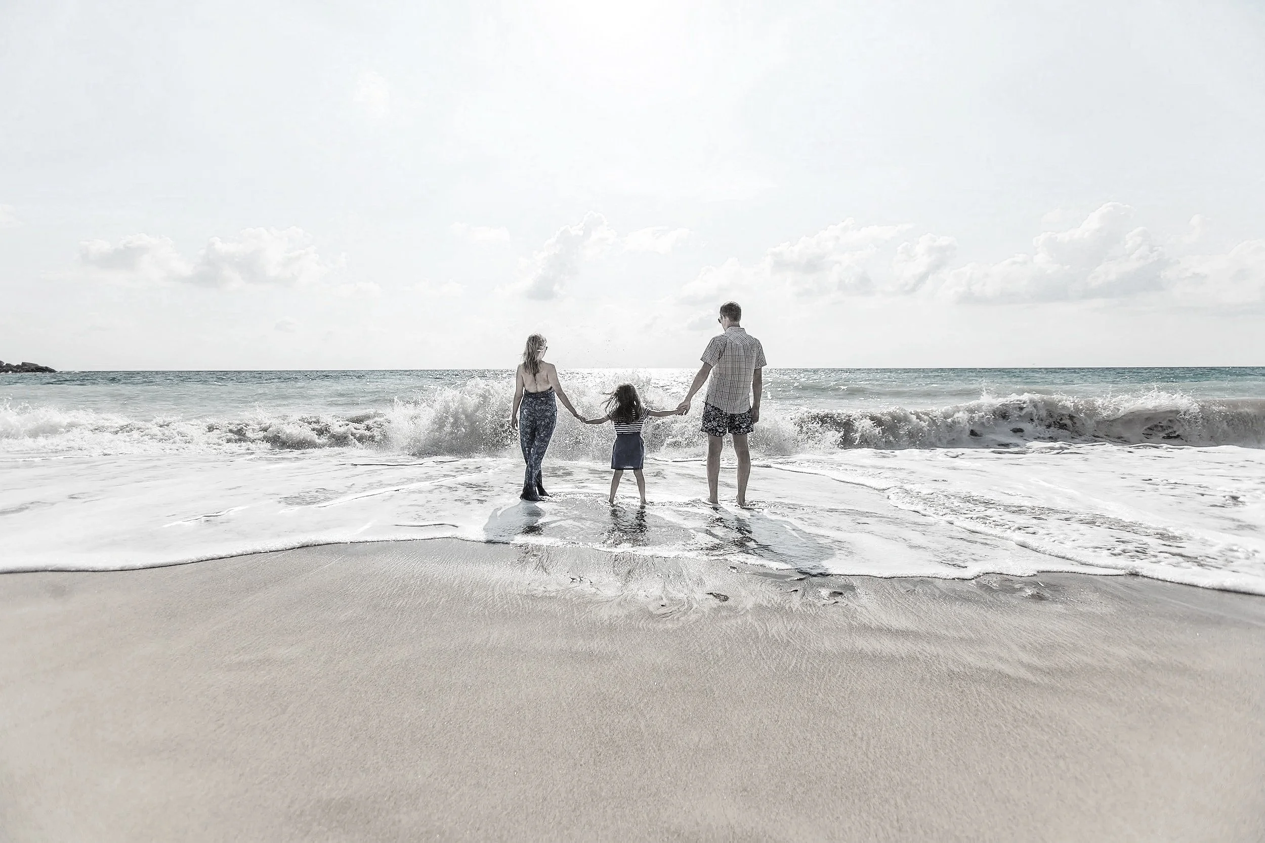 A family of three holding hands at the beach, standing in the shallow water with waves crashing around them, facing the ocean under a partly cloudy sky.