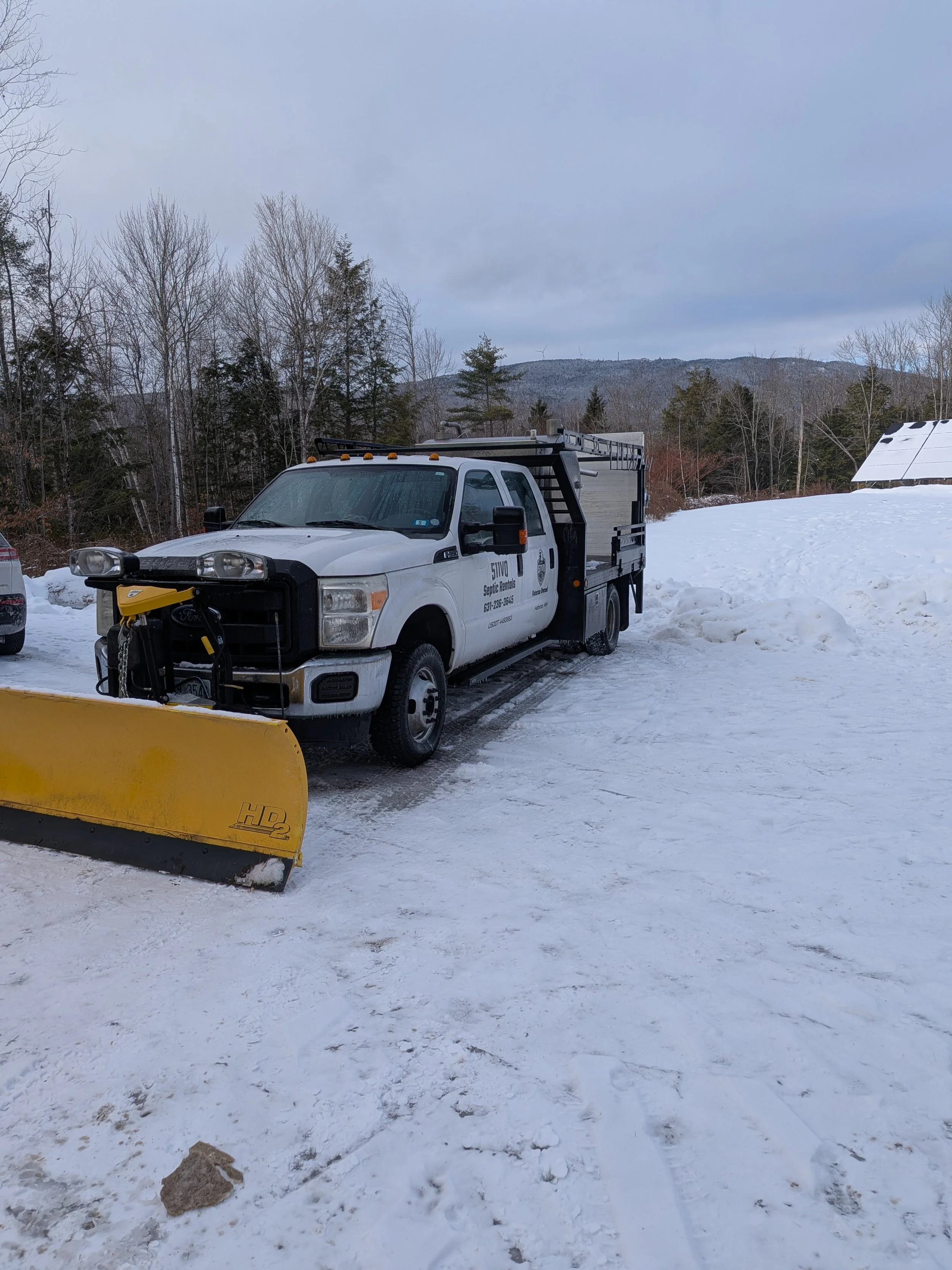 A white service truck equipped with a yellow snow plow attachment parked on a snowy surface, with a landscape of leafless trees and distant hills in the background.