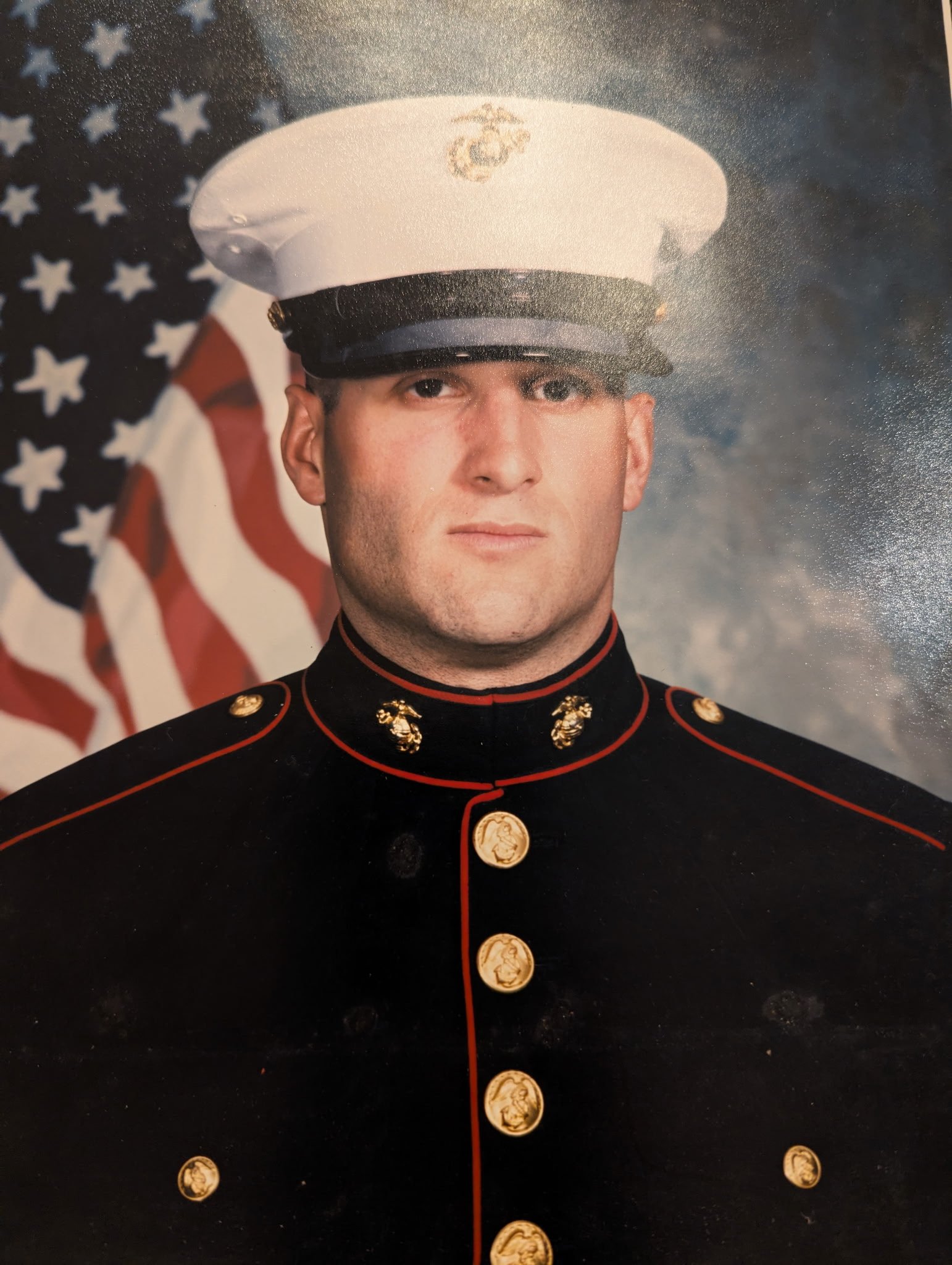 A man in a United States Marine Corps dress uniform with a white hat, standing in front of an American flag.