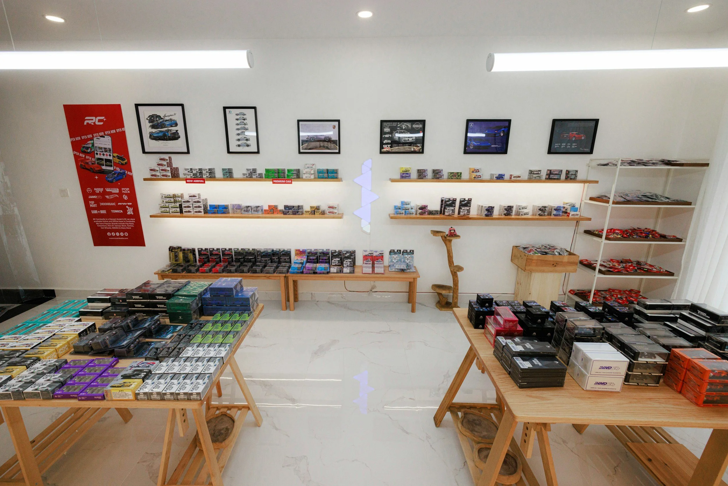 A retail store with wooden tables and shelves displaying various small collectible items, organized in rows and packages, with framed pictures of cars on the white wall, a red promotional banner on the left, and a light-colored marble floor.