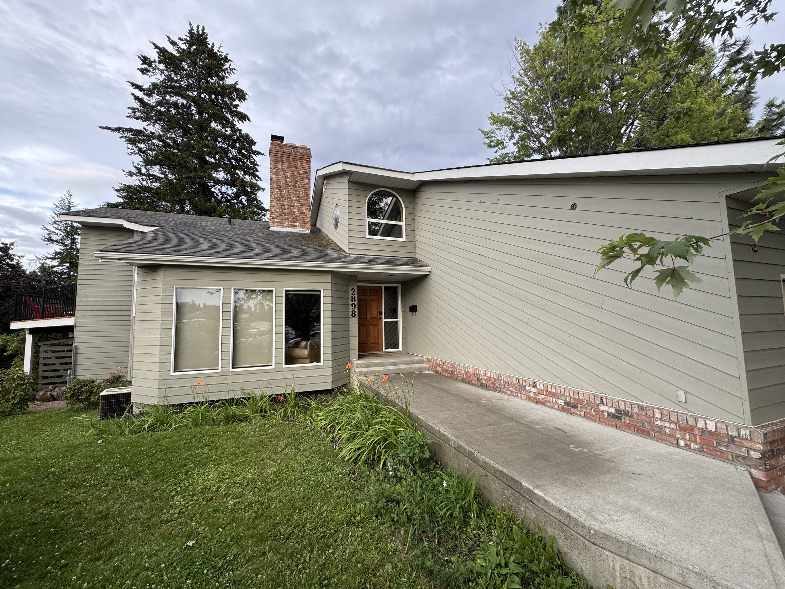 Front of a house with a brick chimney, grey siding, a wooden front door, and large front windows. The house has a small lawn with flowers and a concrete walkway leading to the door. There are trees in the background and a cloudy sky overhead.