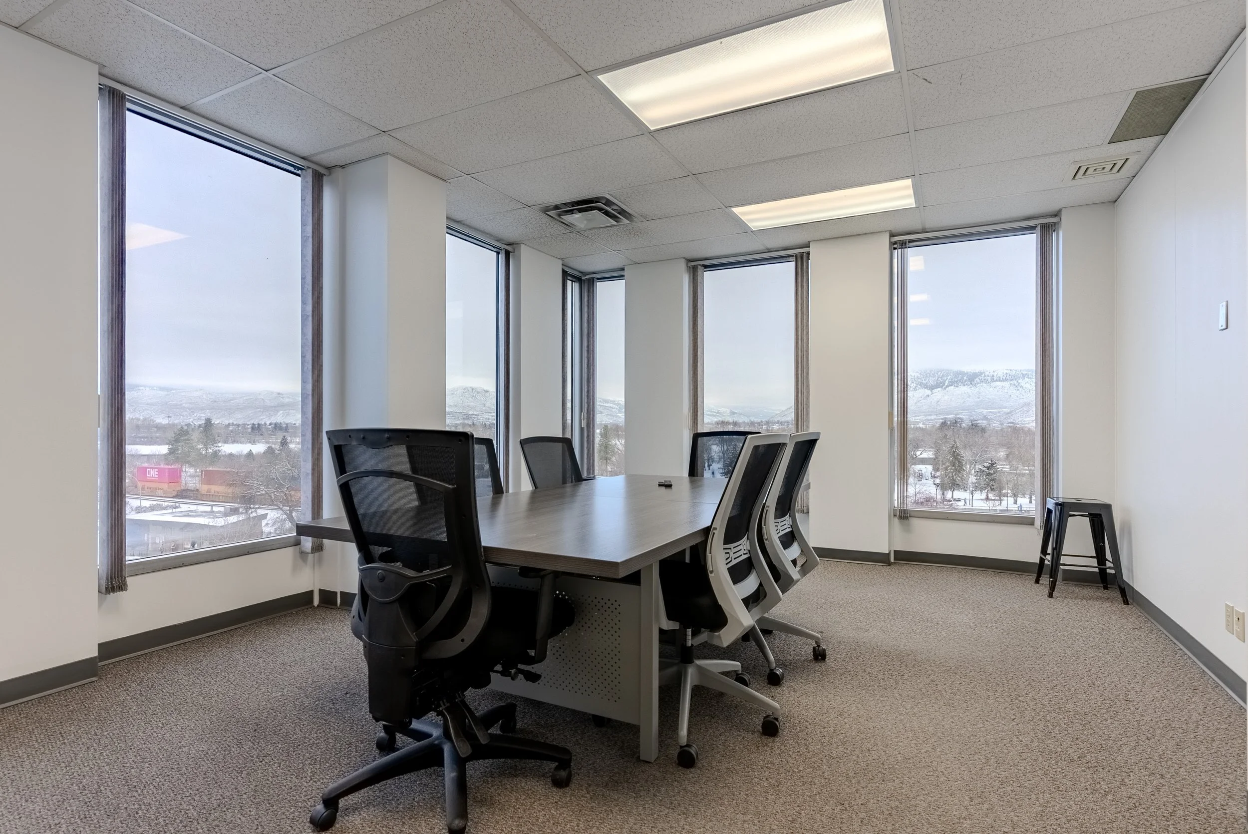 An empty conference room with a wooden table and six office chairs, five on one side and one on the other, surrounded by large windows showing a snowy landscape outside.