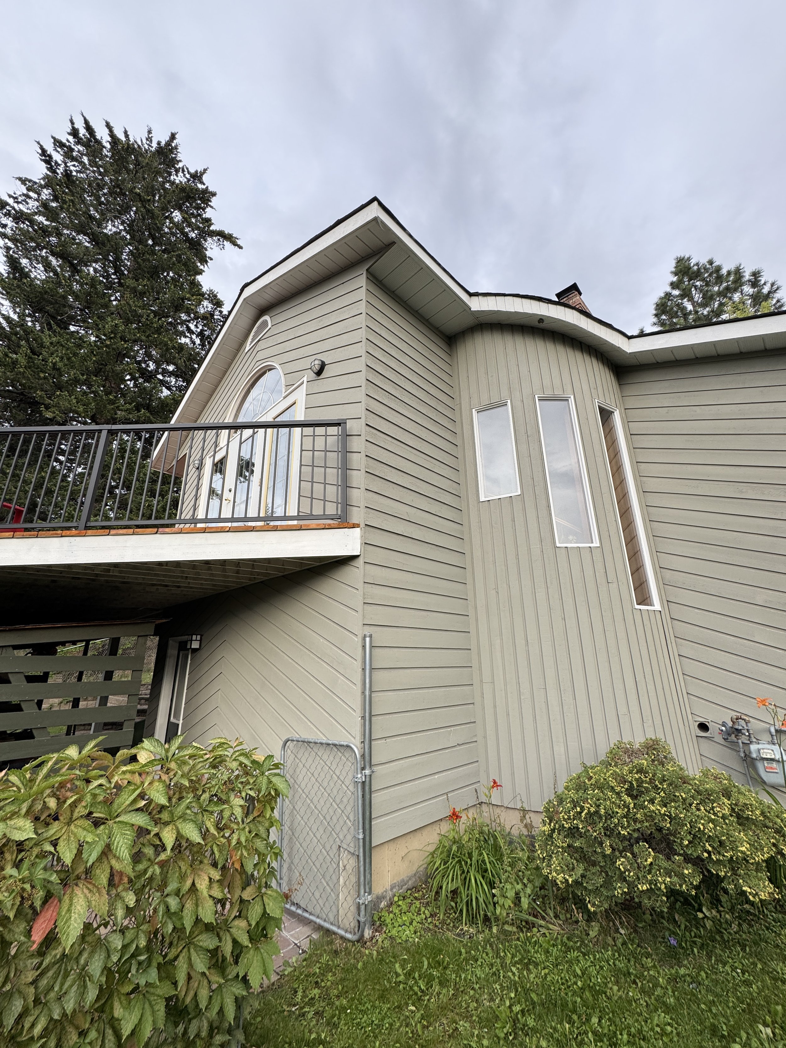 A multi-story house with beige wooden siding, multiple tall, narrow windows, a balcony with black metal railing, and trees in the background under a cloudy sky.