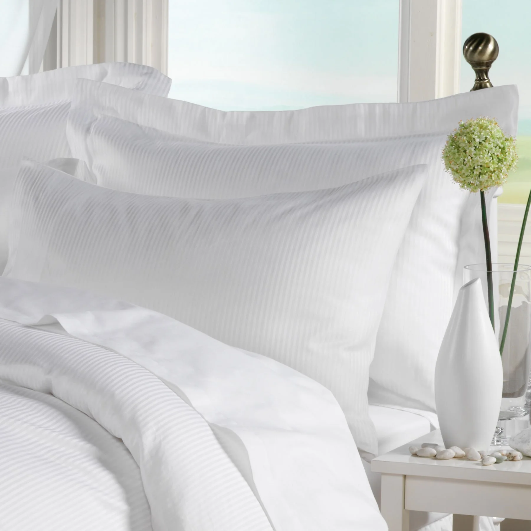 A neatly made bed with white striped bedding and pillows, a white bedside table with white stones, a white vase with a green flower, and a window in the background.