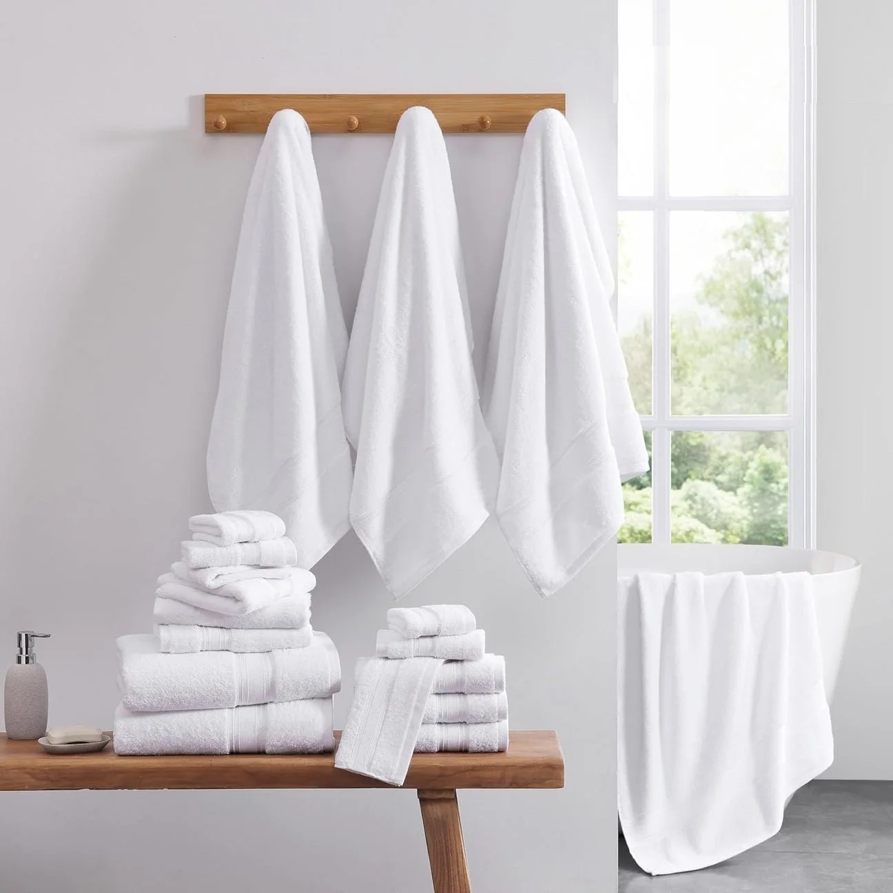 White towels hanging on a wooden rack, with stacked towels on a table in front, and a bathtub draped with a towel next to a large window with green trees outside.