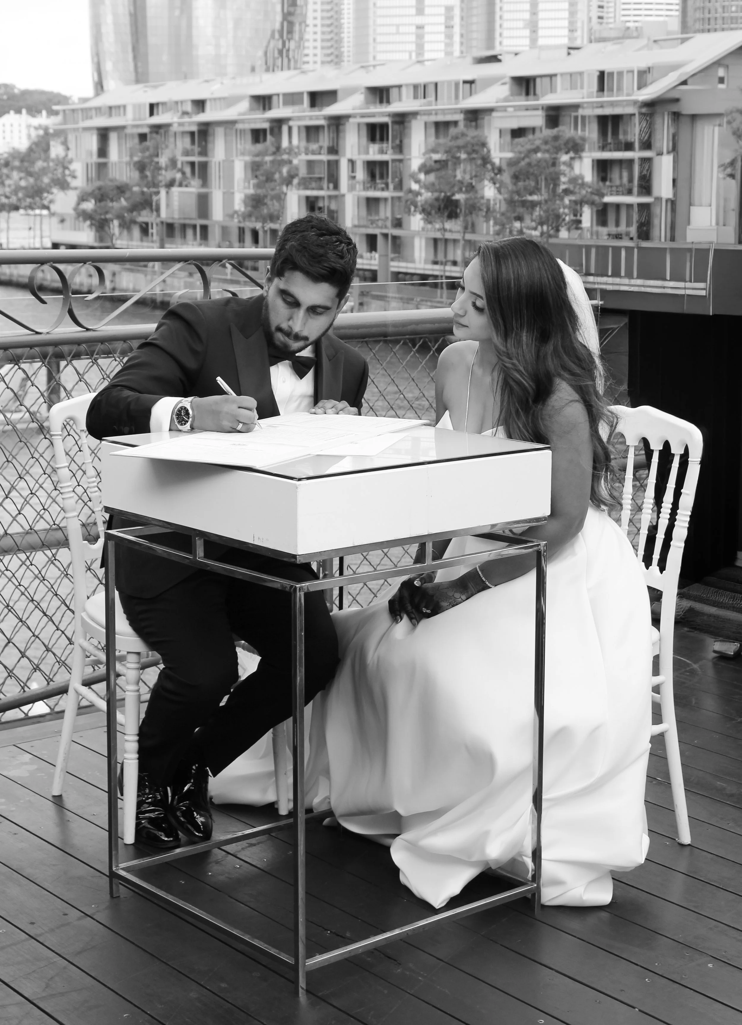 A black-and-white photo of a man and woman sitting at a small table on an outdoor balcony. The man, dressed in a tuxedo, is signing a document, while the woman, wearing a wedding dress, watches. The background shows a cityscape with modern apartment buildings and trees.